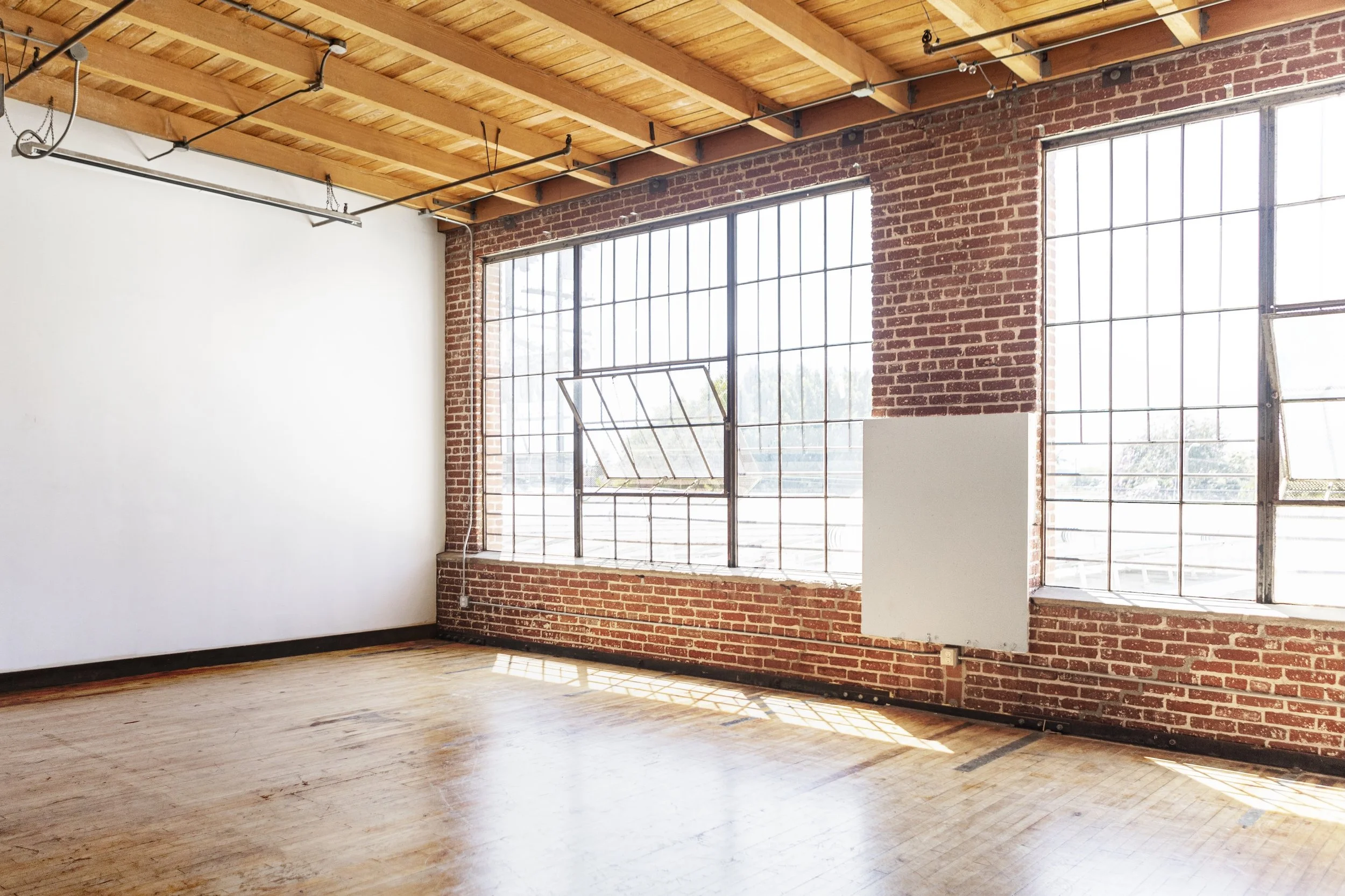 An empty room with wooden flooring, exposed brick walls, and large industrial-style windows allowing natural light to fill the space.