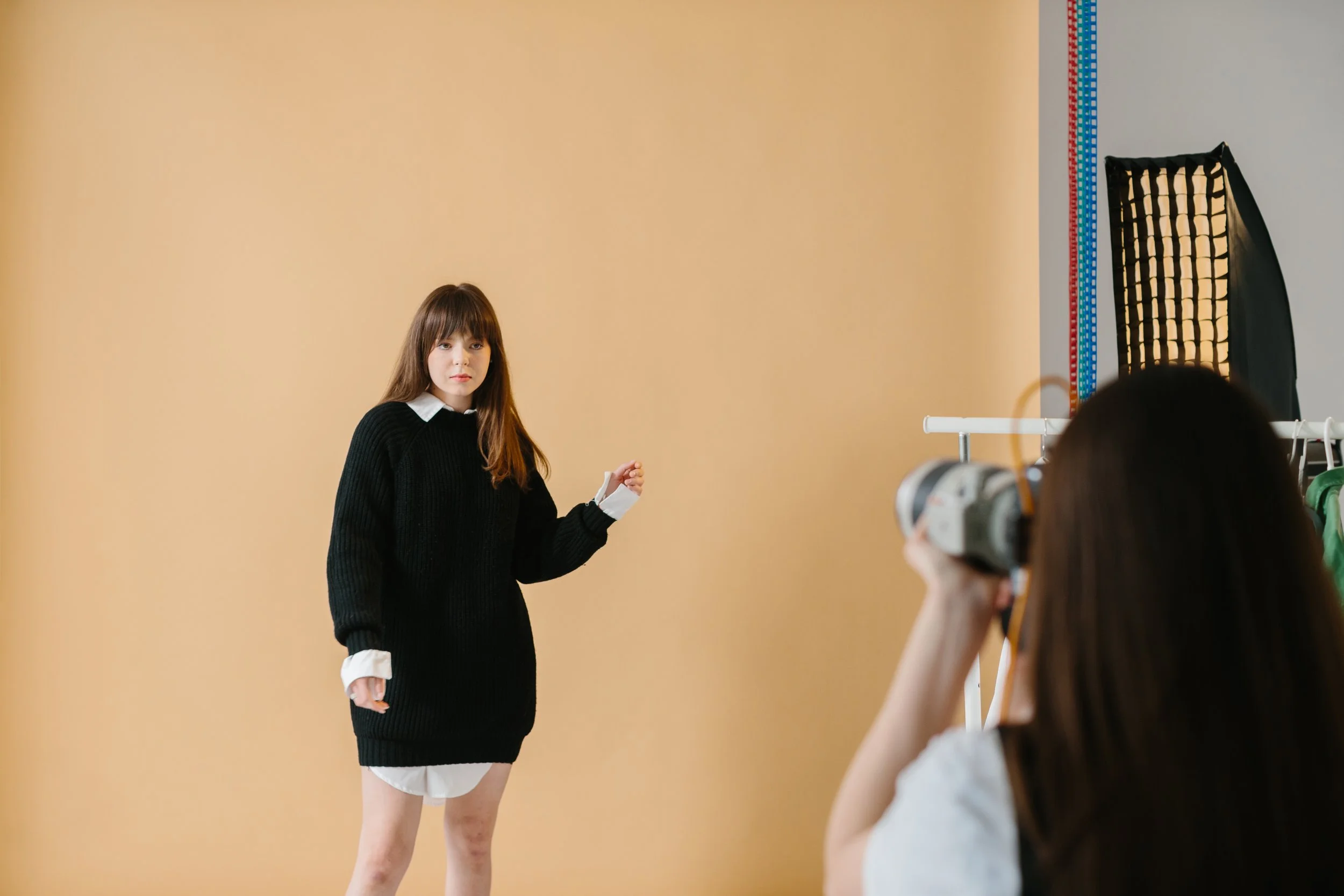 A young woman with long brown hair stands against a beige wall during a photoshoot, wearing a black sweater and a white shirt underneath. She is looking at the camera with a serious expression. A photographer with dark hair, wearing glasses, is visible in the foreground holding a camera. Studio lighting equipment is visible to the right of the image.