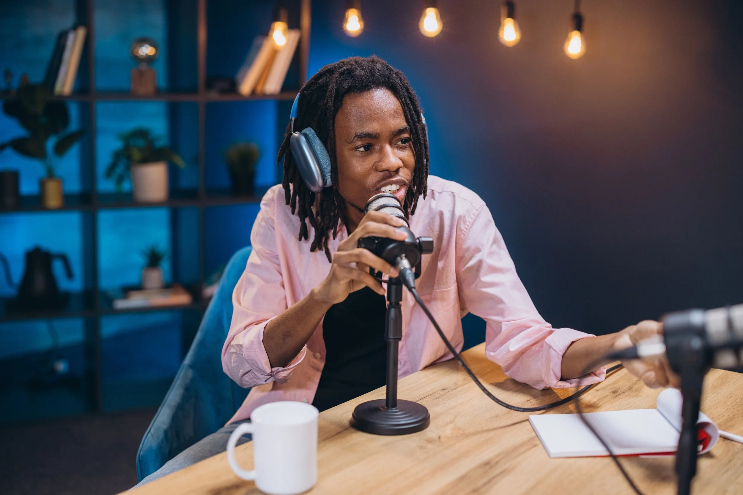 A person with dreadlocks wearing headphones and a pink shirt sitting at a table, speaking into a microphone with a studio background decorated with shelves, plants, notebooks, and warm hanging lights.