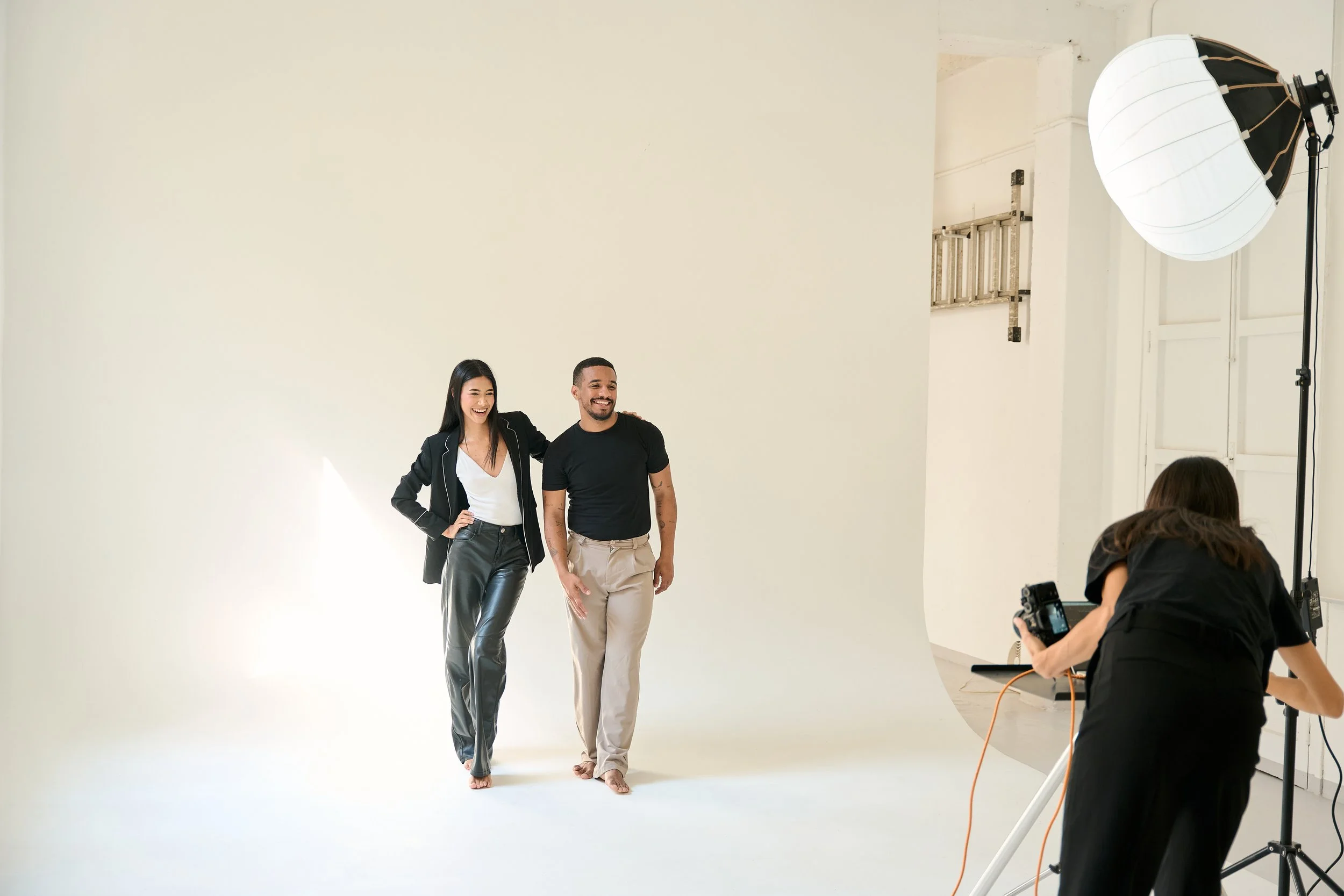 Two models, a woman and a man, smiling and walking barefoot in a studio with a white backdrop during a photo shoot. The woman has long black hair and is wearing a black blazer and shiny black pants, while the man has short hair and a beard, wearing a black T-shirt and beige pants. A photographer is taking their picture, and a large studio light with a softbox is visible on the right side.