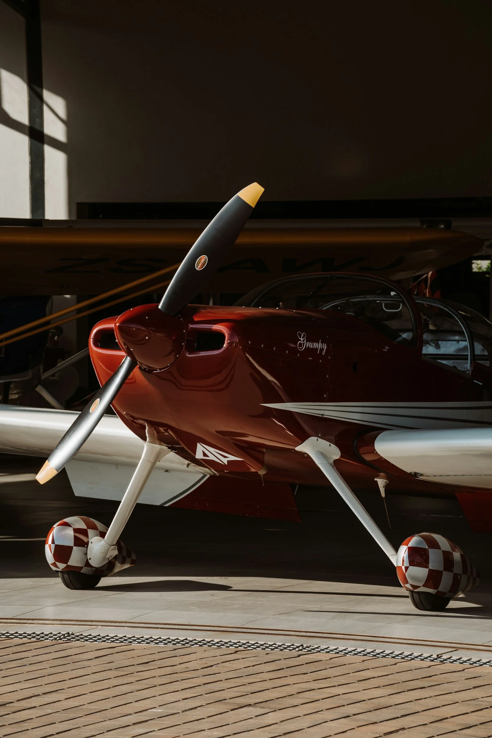 Side view of a small, red, single-engine airplane with checkered wheels parked on a tarmac, partially shaded, with a large flat surface behind it.