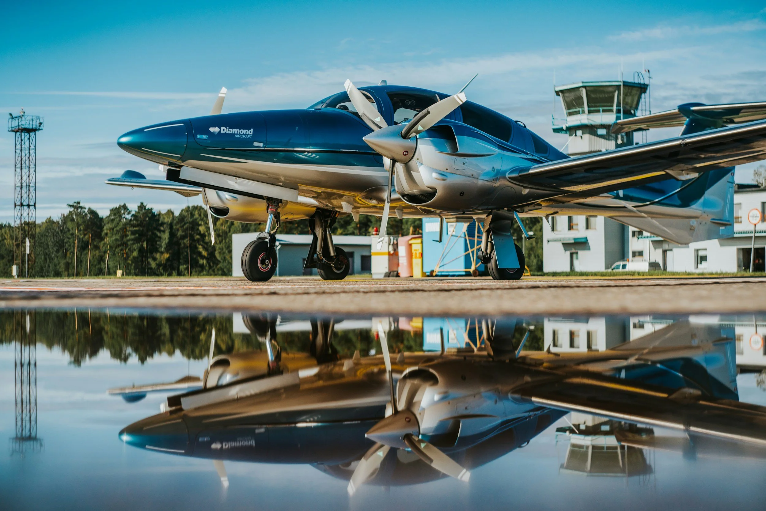 A sleek, futuristic aircraft parked on an airport tarmac with trees and airport buildings in the background, reflected in a puddle of water below.