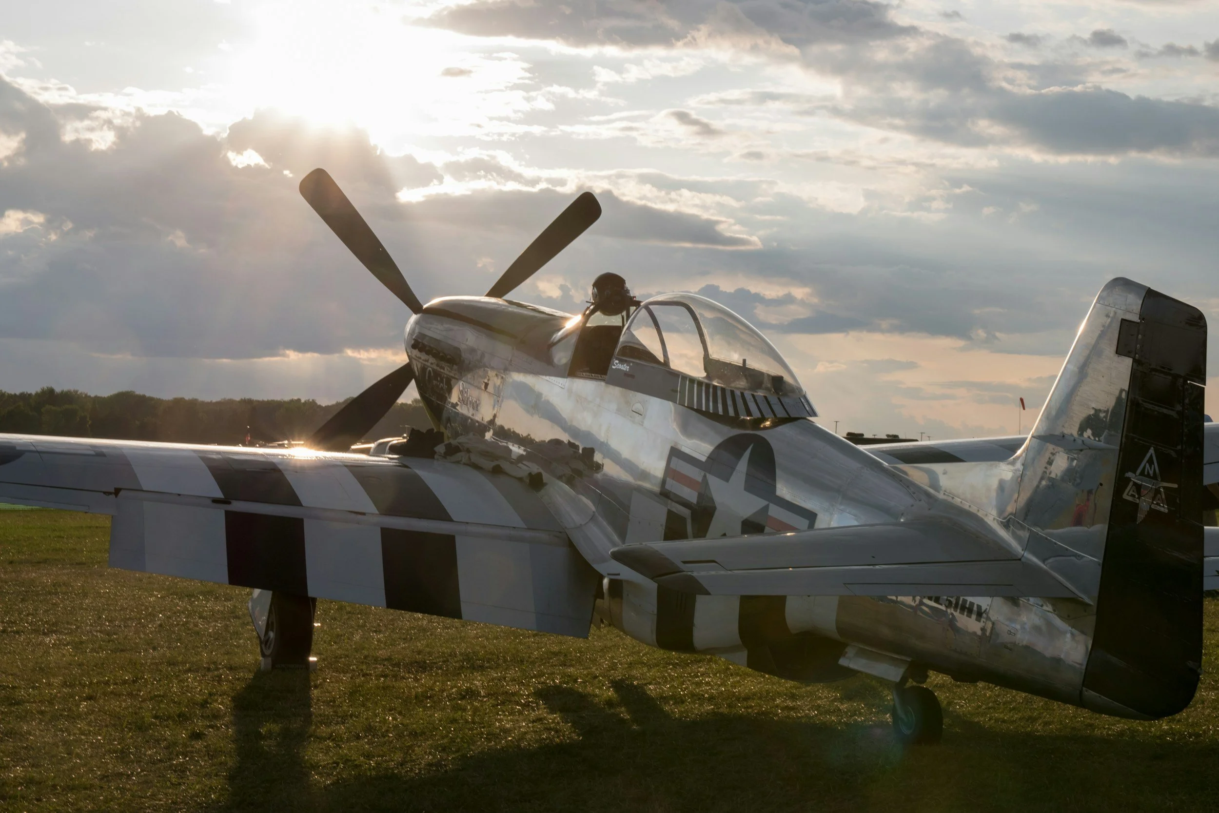 A vintage fighter plane parked on grass with the sun setting or rising behind it, casting a glow on the aircraft and creating a partly cloudy sky.