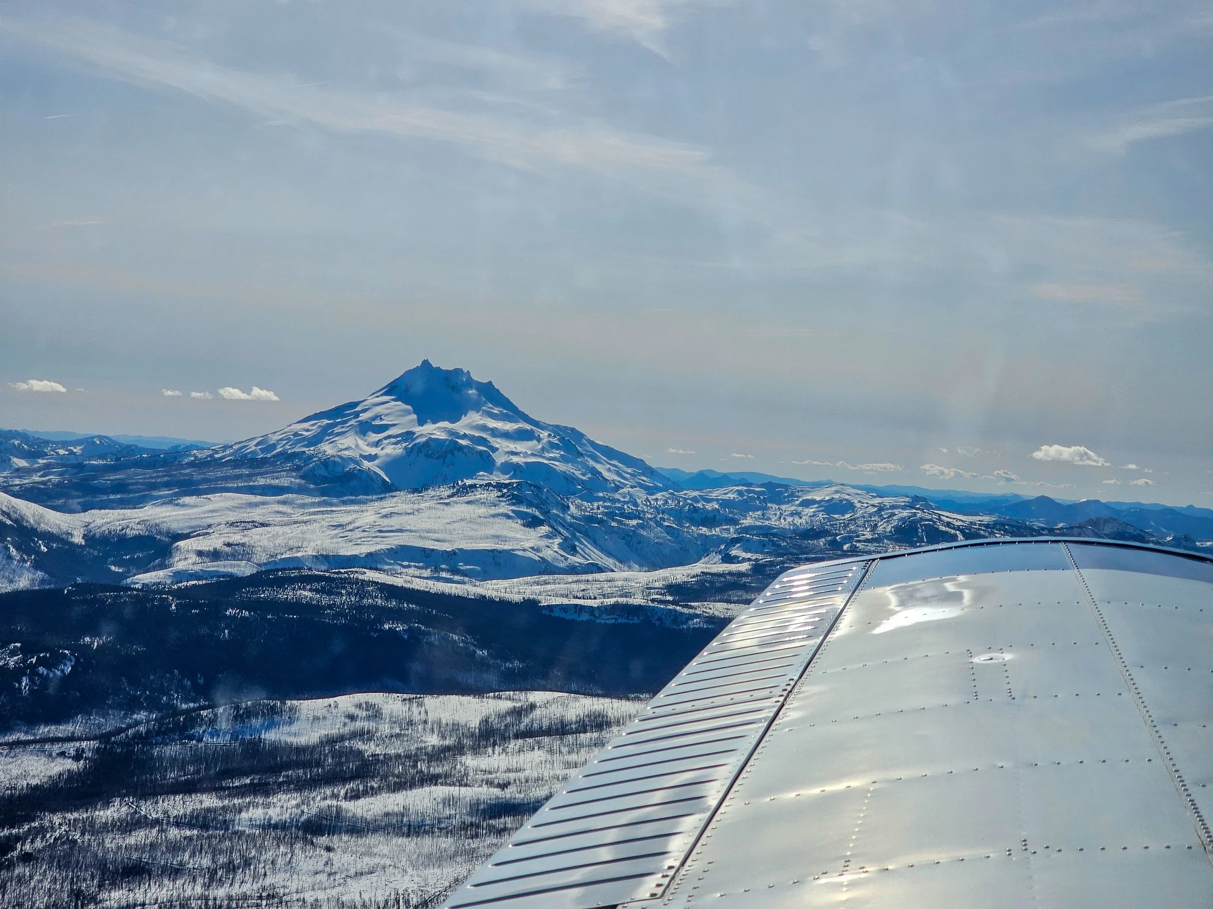 Snow-covered mountain, likely Mount Hood, with a partly cloudy sky and airplane wing visible in the foreground.
