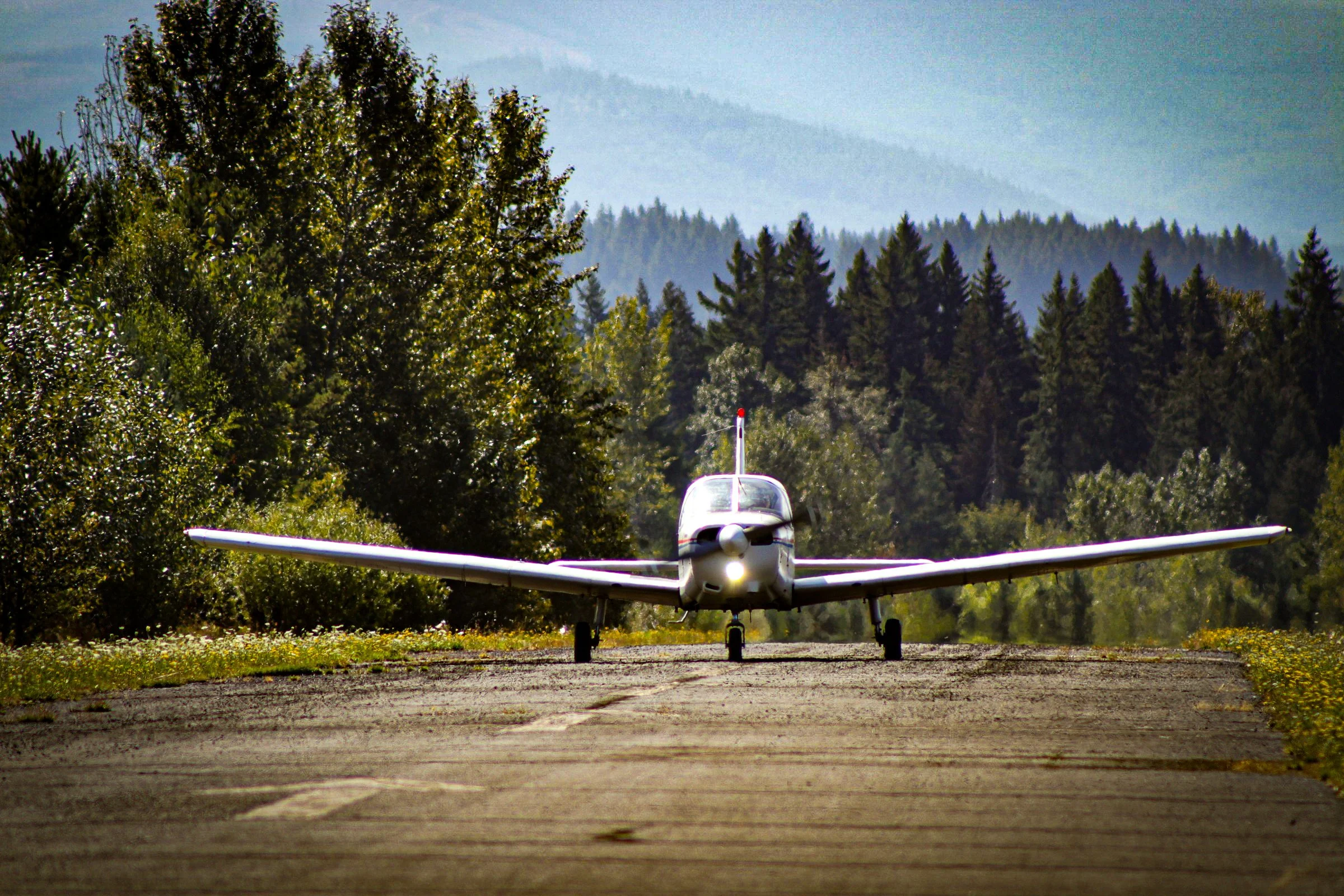 Small white airplane on a grassy airstrip surrounded by trees with mountains in the background.