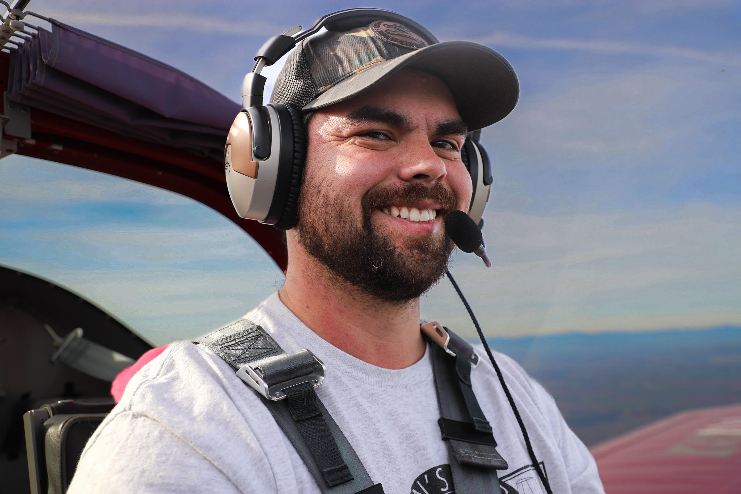 A smiling man with a beard wearing headphones, a camouflage hat, and a white t-shirt, standing outdoors near an aircraft wing with a blue sky in the background.