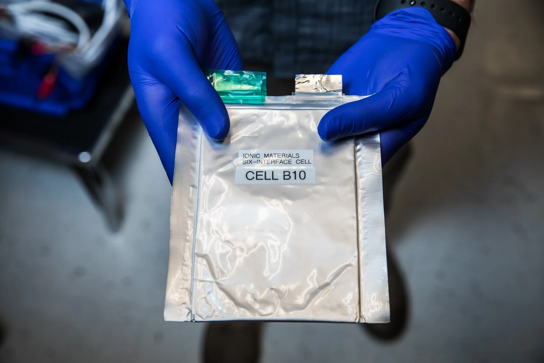 Person wearing blue gloves holding a sealed silver bag labeled 'IONIC MATERIALS SIX-INTERFACE CELL CELL B10' in a laboratory setting.