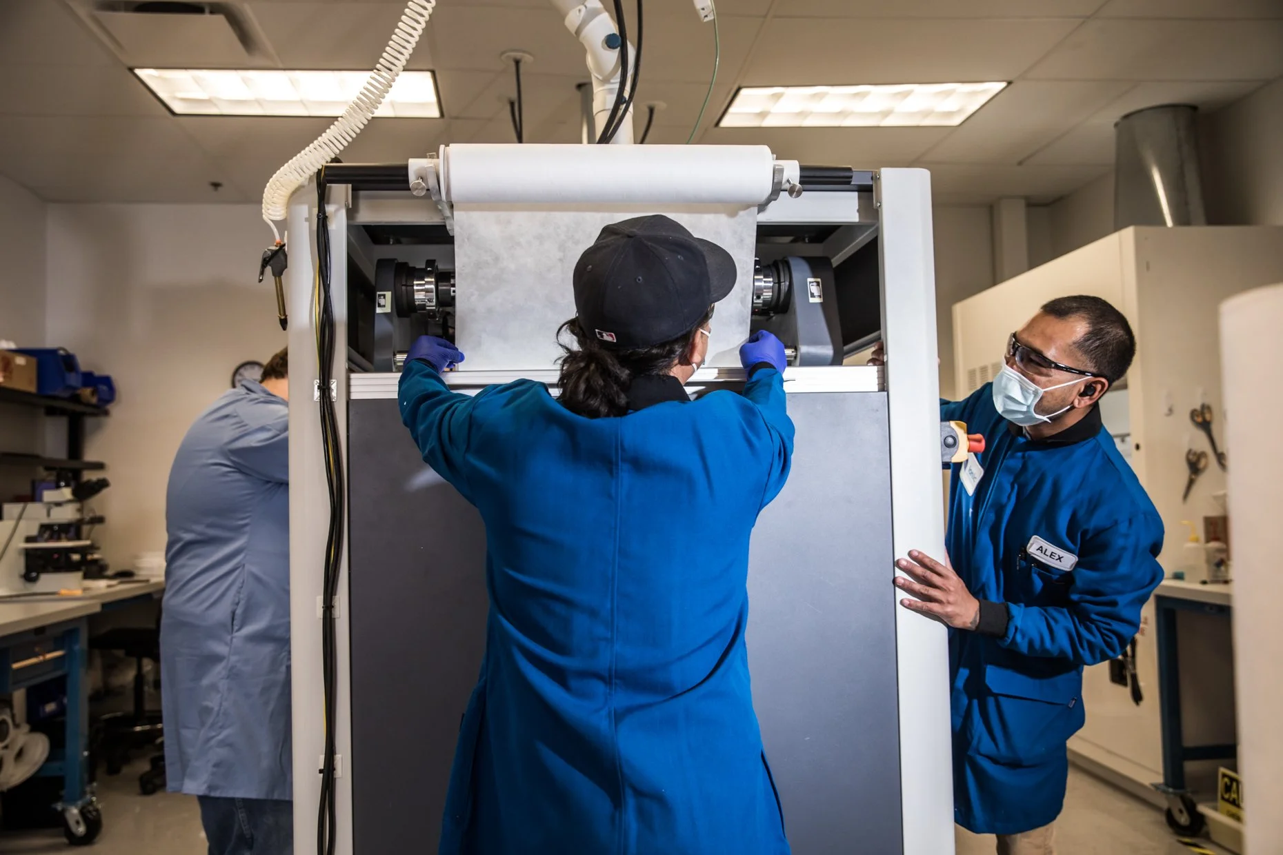 Scientists working with a large scientific machine in a lab. The person in the center, wearing a black cap and blue lab coat, is adjusting the machine. The person on the right, with a name tag reading 'ALEX' and wearing safety glasses and a face mask