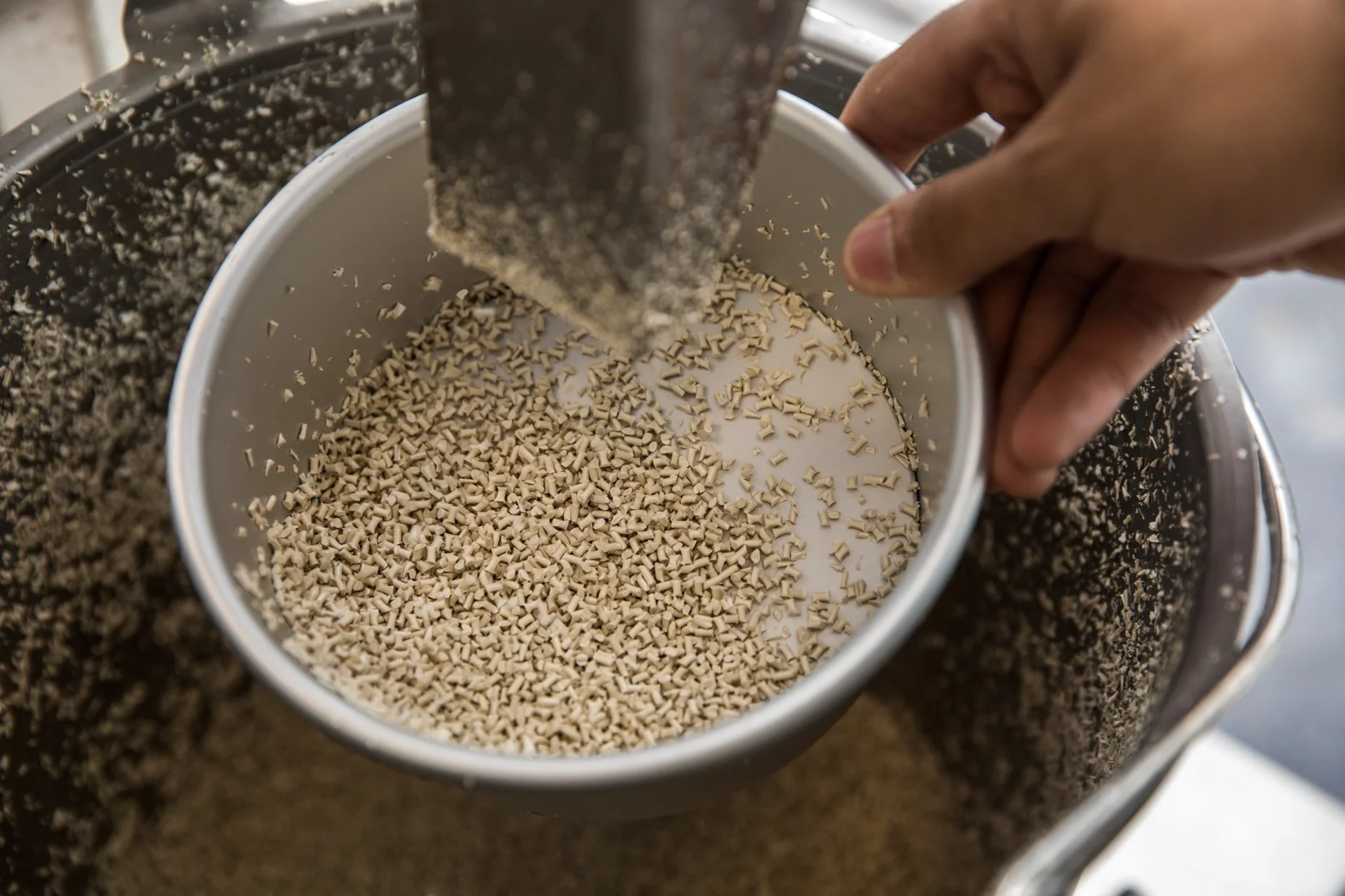 Close-up of a person's hand pouring tiny beige pellets into a white container, with a dark chocolate-colored container nearby filled with more of the pellets.