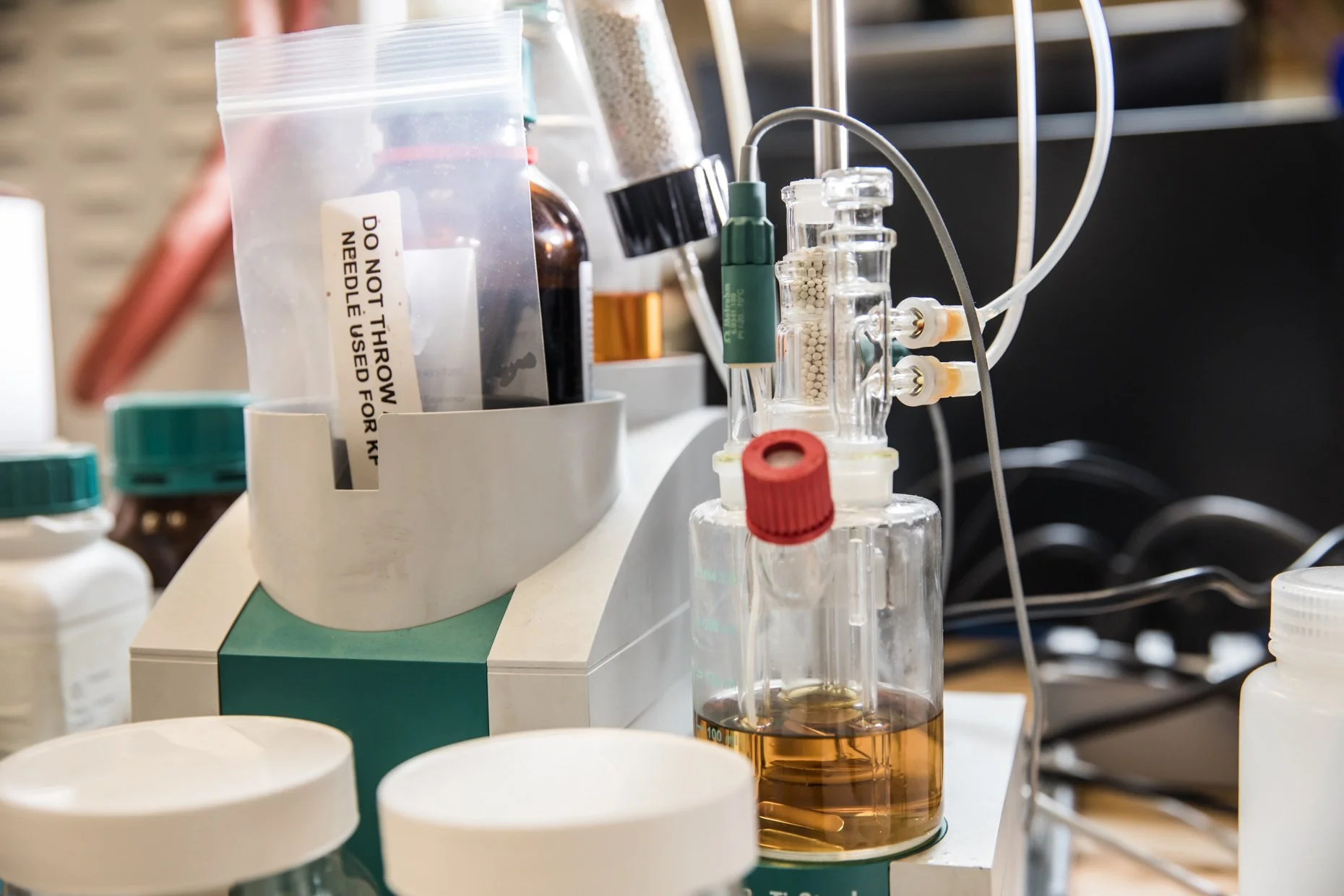 Lab workspace with clear glass containers, some with brownish liquids, connected with tubes and equipment, along with bottles and containers in the background.