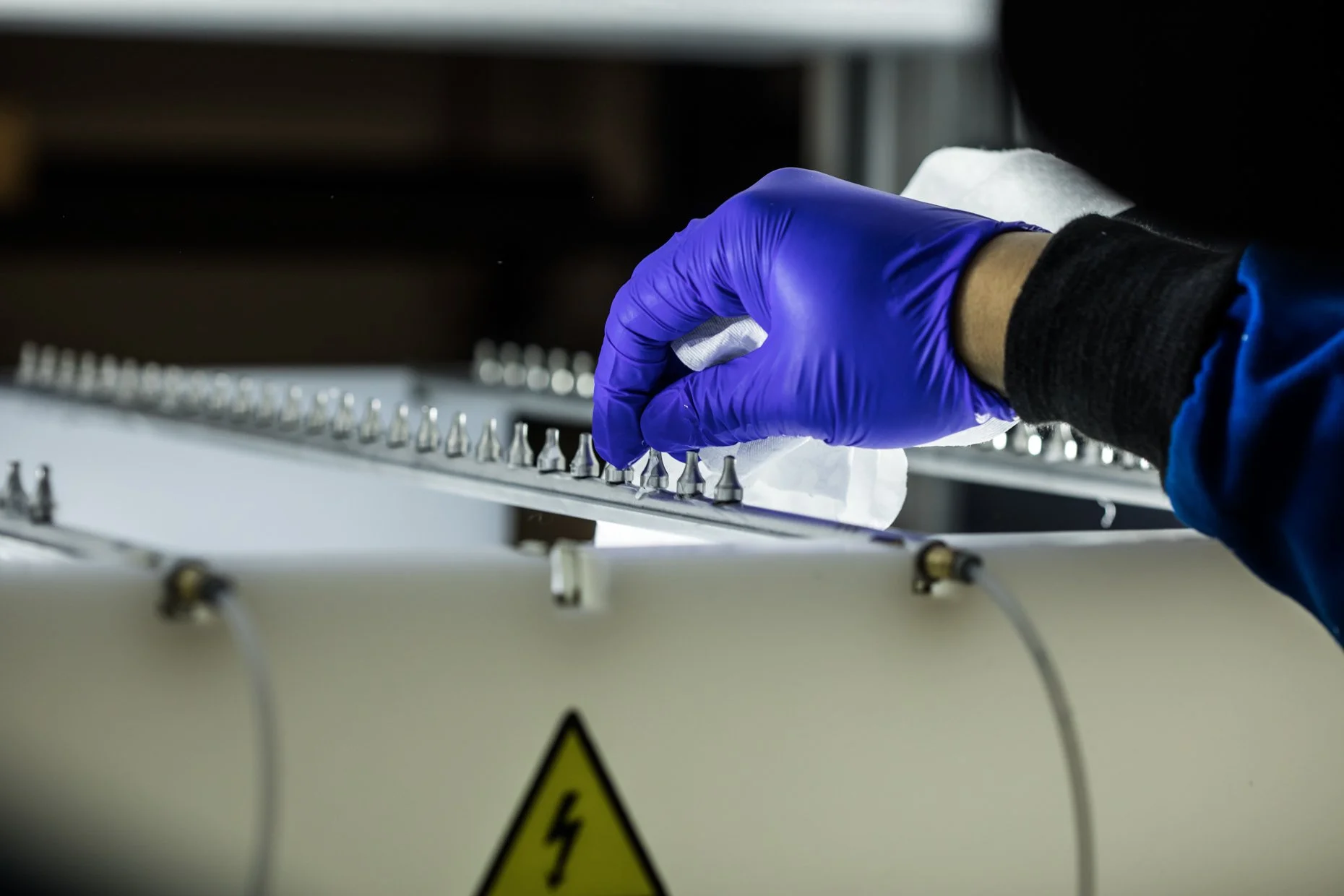 A person wearing a blue sleeve and purple glove cleaning or inspecting an electrical device with multiple small components, possibly in a laboratory or industrial setting.
