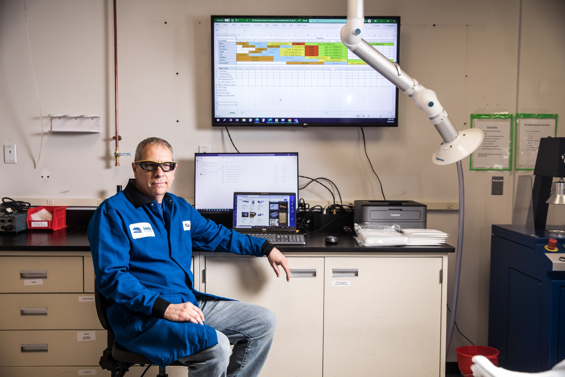 A man wearing protective glasses and a blue work uniform sitting in a laboratory or medical setting, with computer monitors and lab equipment on the desk behind him.
