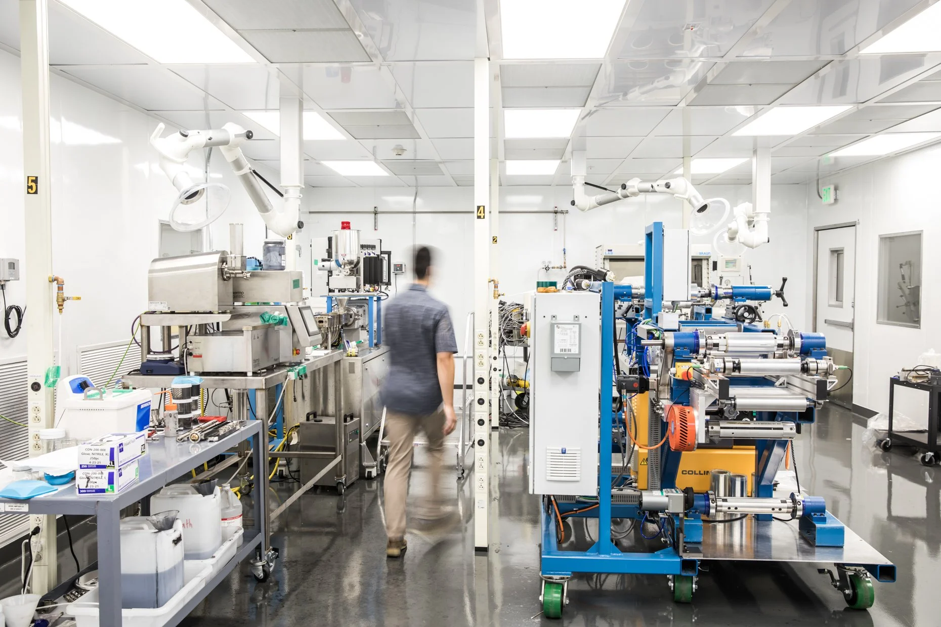 A researcher working in a laboratory with scientific equipment including robotic arms and machinery, with a man walking between the equipment.