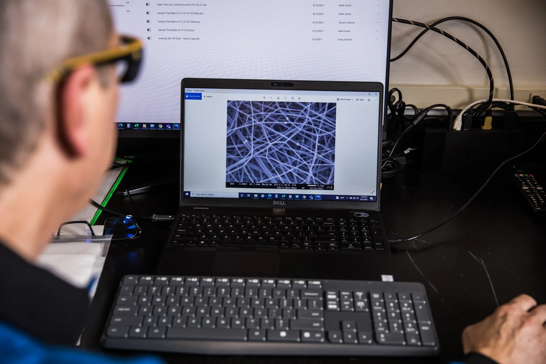A person with glasses working at a desk with two monitor screens. The smaller screen in front of the person displays a microscopic image of tangled fibers, while the larger monitor behind shows a list of Excel file names and dates.