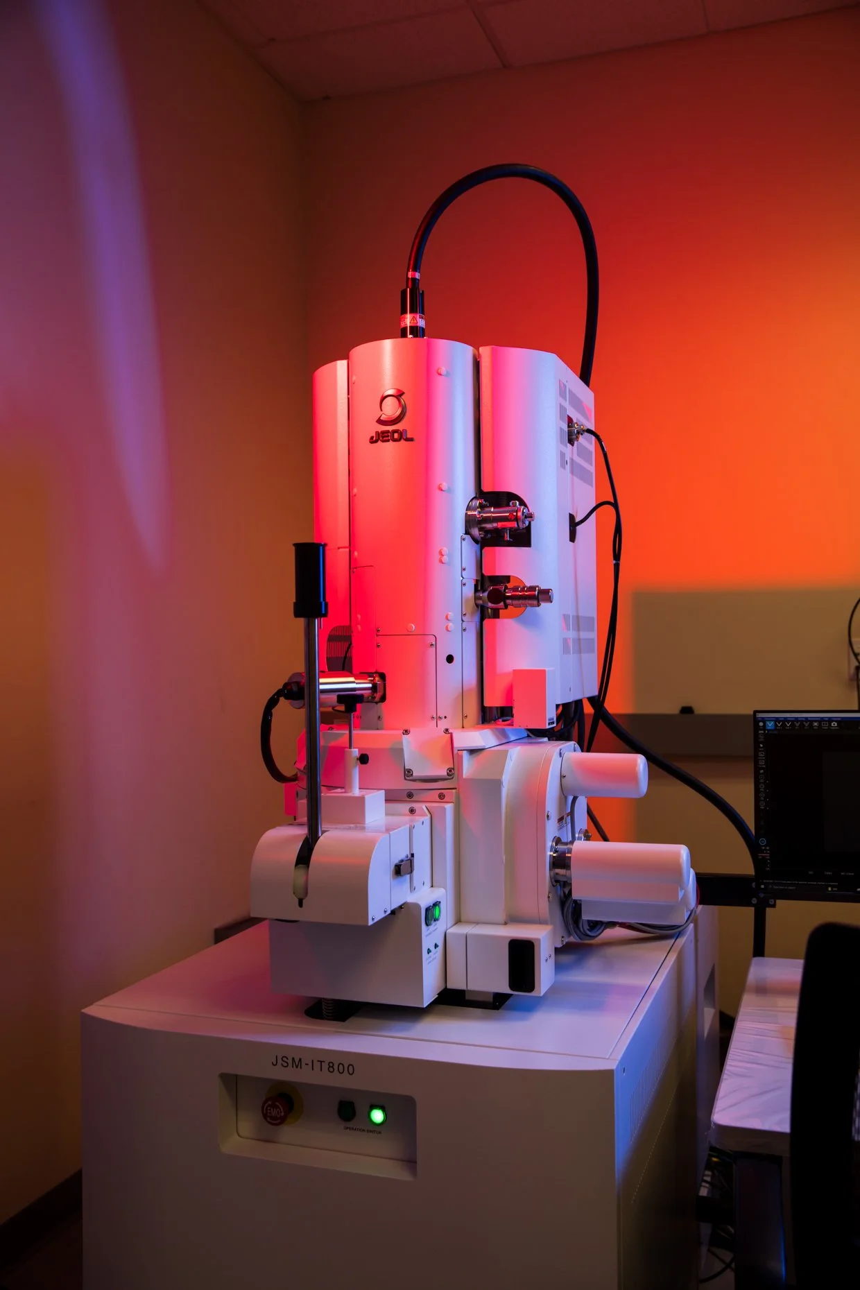Laboratory microscope with white body and black cables, illuminated by red and purple lighting, placed on a table with a monitor nearby.