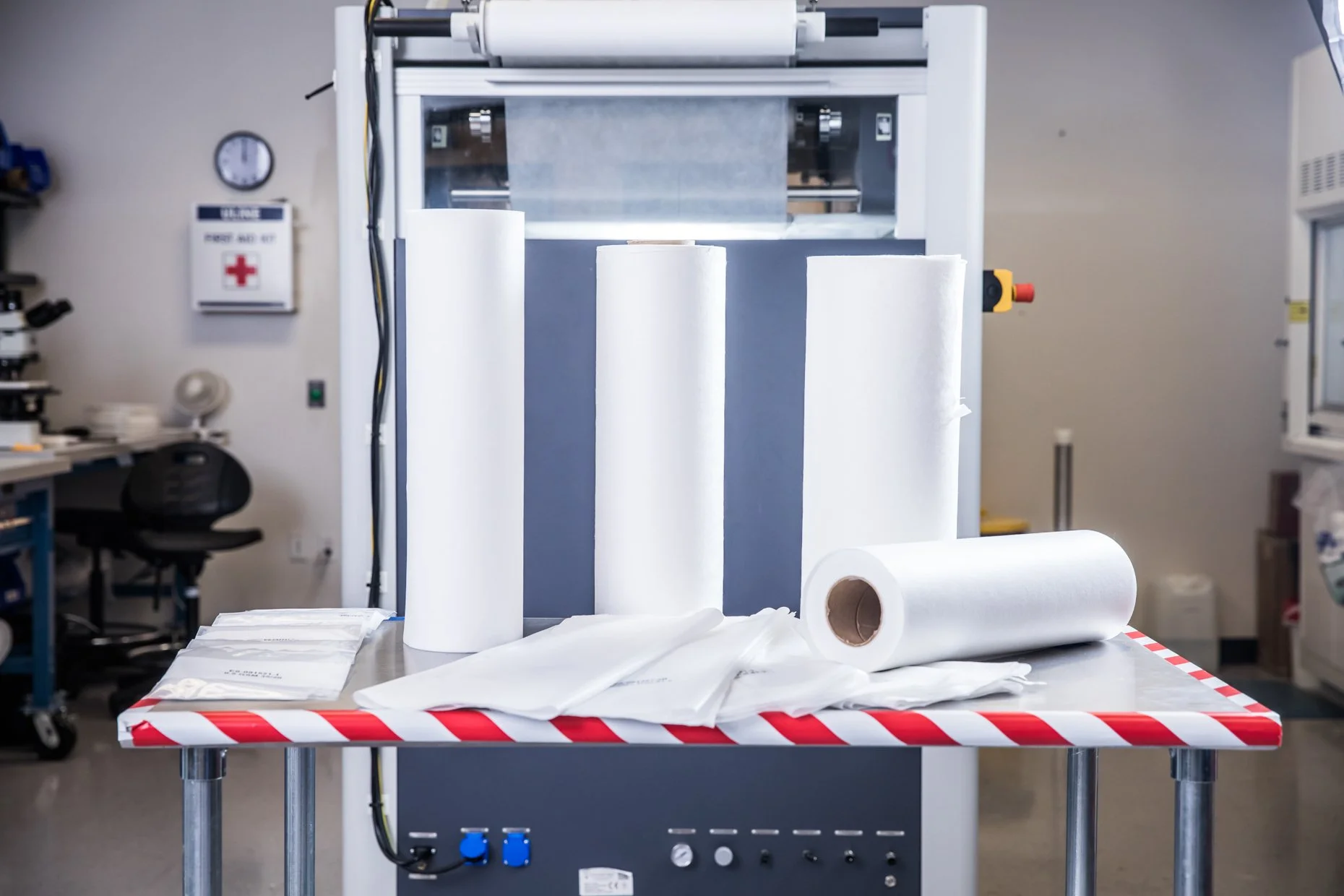 A large industrial machine in a laboratory or manufacturing setting with rolls of paper towels or similar material on a worktable in front.