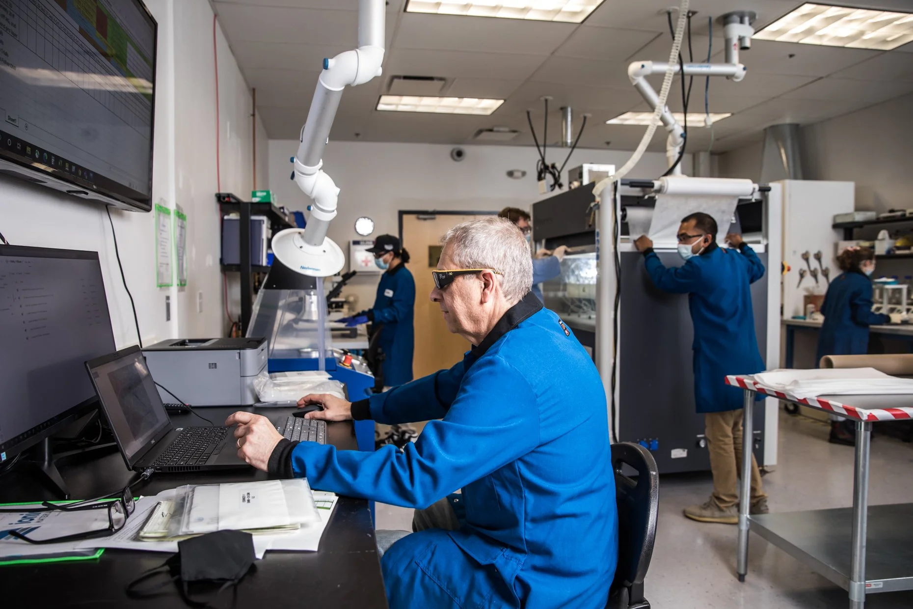 Researchers in a laboratory working with specialized equipment and computers, wearing blue lab coats and face masks.