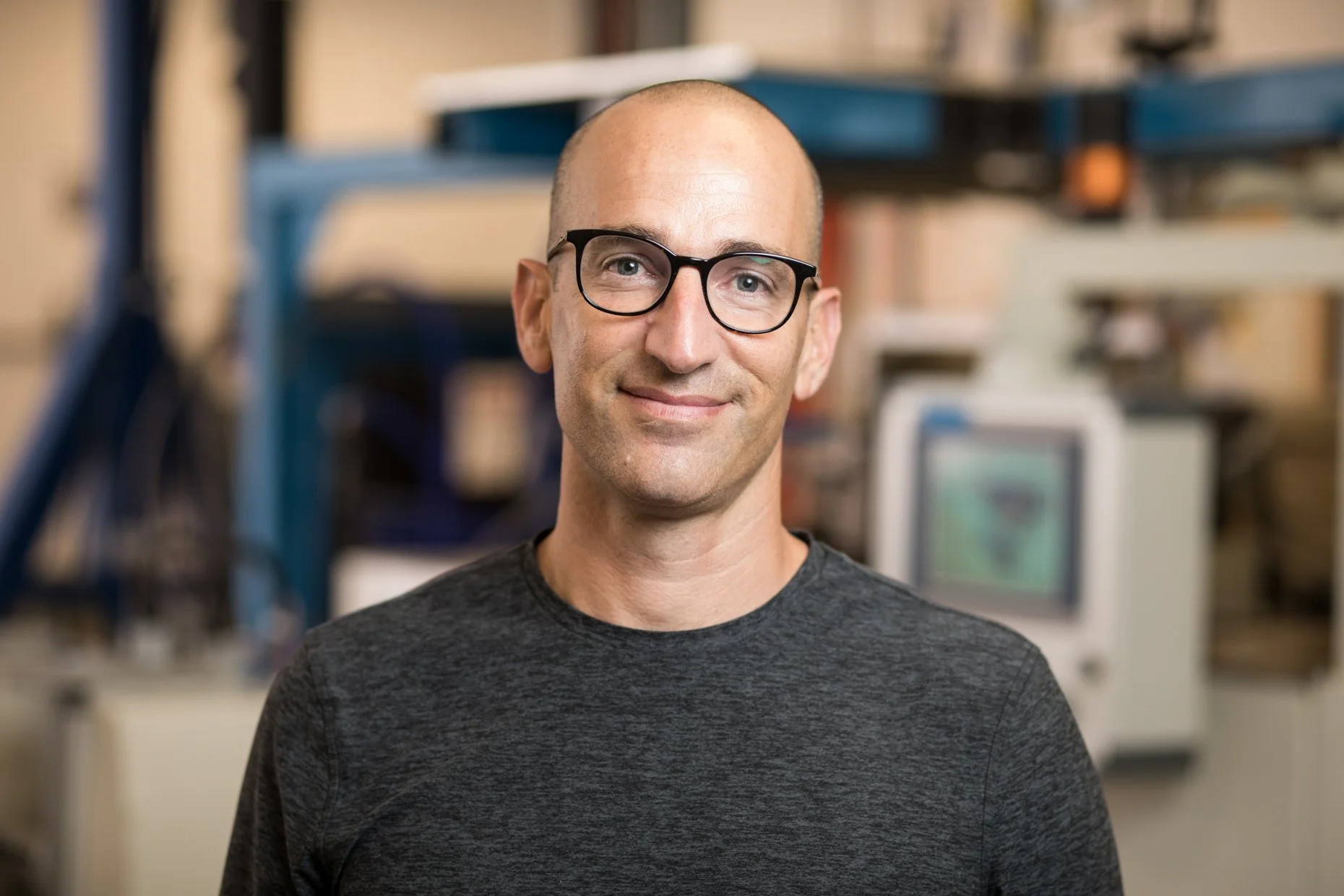 A man with a shaved head and glasses smiling in a workspace with shelves and computer equipment in the background.