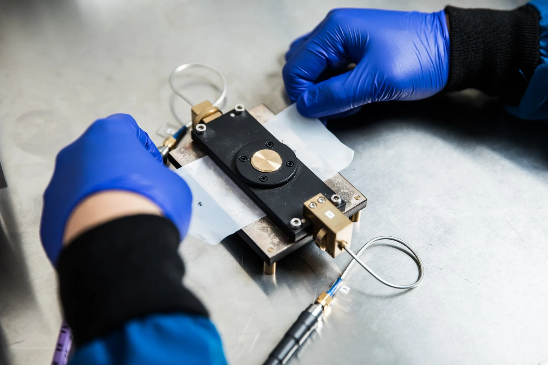 Laboratory technician wearing blue gloves assembling scientific equipment with a black and gold component on a metal surface.