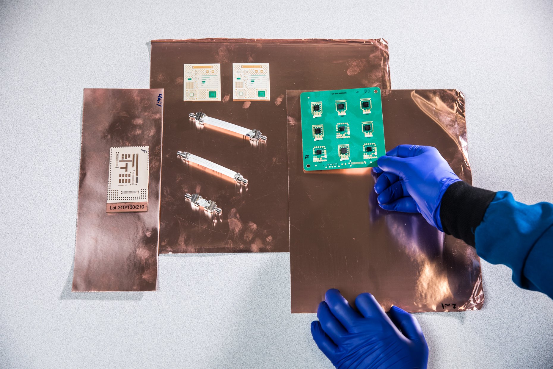 A person wearing blue gloves holding a green circuit board with black chips on a copper work surface. There are other electronic components and printed circuit boards on the table.