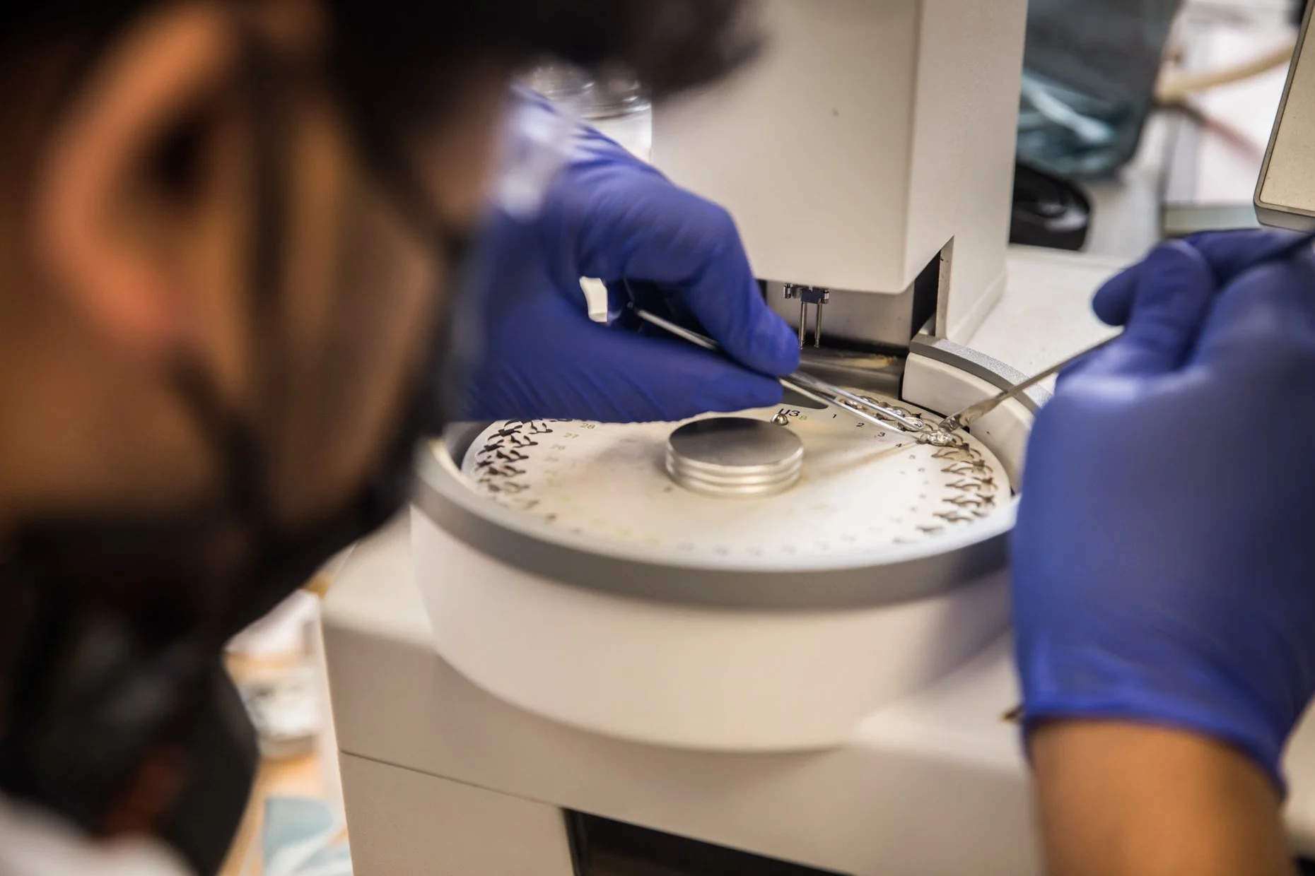 A scientist wearing blue gloves operates a laboratory machine with a circular rotor and metallic tools, likely involving sample preparation or analysis, in a scientific lab setting.