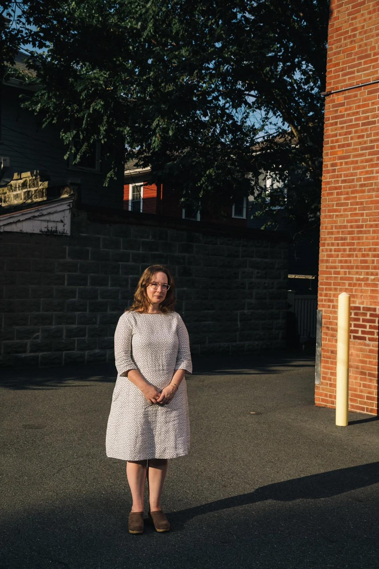 A woman with brown hair and glasses wearing a white dress with black polka dots stands outdoors on a paved surface in the evening sunlight, with a brick wall, a stone wall, and a large tree in the background.