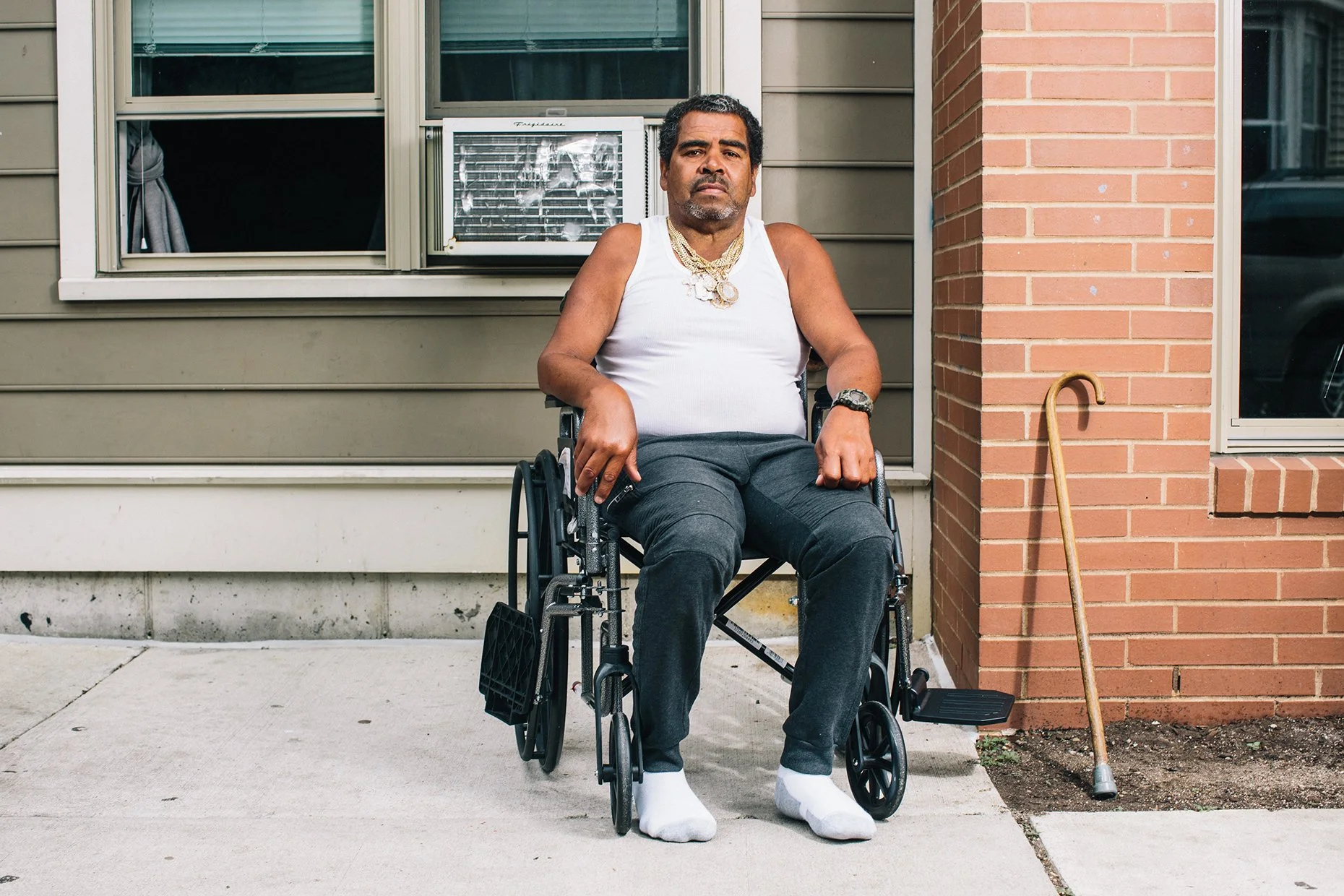 A man in a wheelchair sitting outside a house on a sidewalk, wearing a white tank top, gray pants, white socks, jewelry, a watch, and looking serious. A cane leans against the house on his right side.