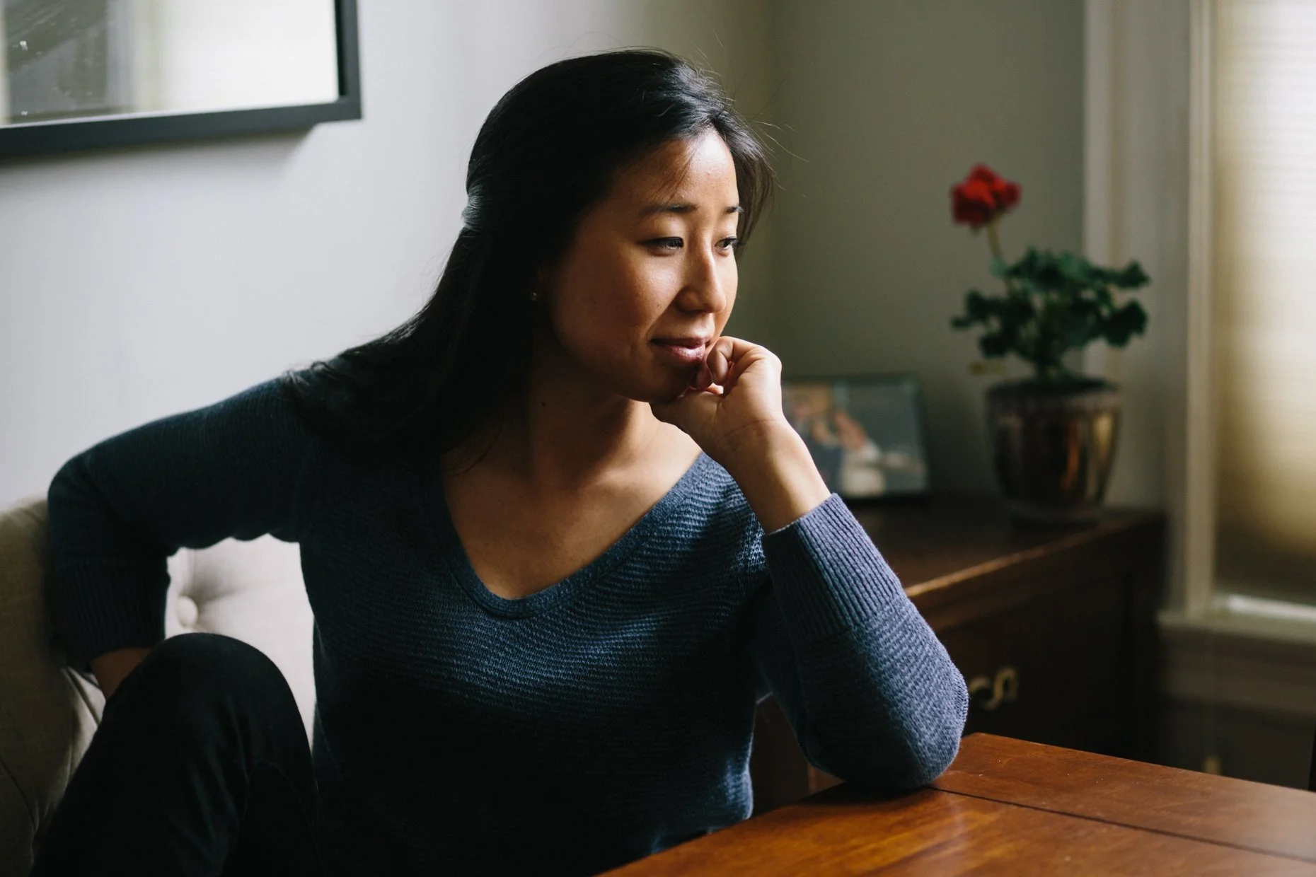 A woman sitting at a wooden table inside a room, looking contemplative with her chin resting on her hand. There is a framed picture and a potted plant with a red flower on a side table in the background.