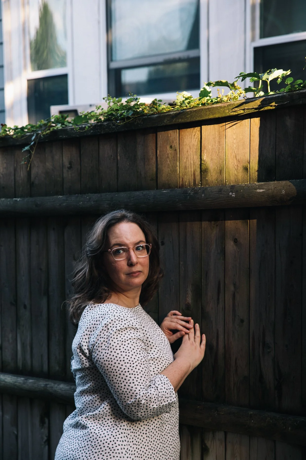 A woman with glasses and wavy brown hair stands outdoors, touching a dark wooden fence with her hands, wearing a white dress with black polka dots.
