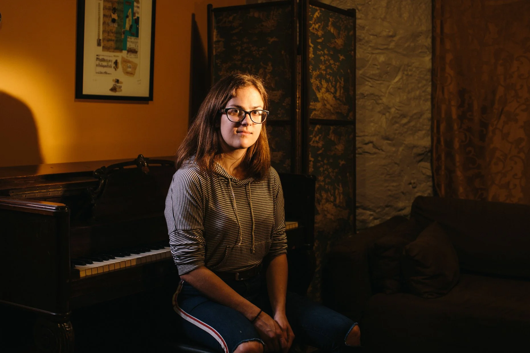 A young woman with glasses and long brown hair sitting on a piano bench in a dimly lit room with a piano and decorative screens in the background.