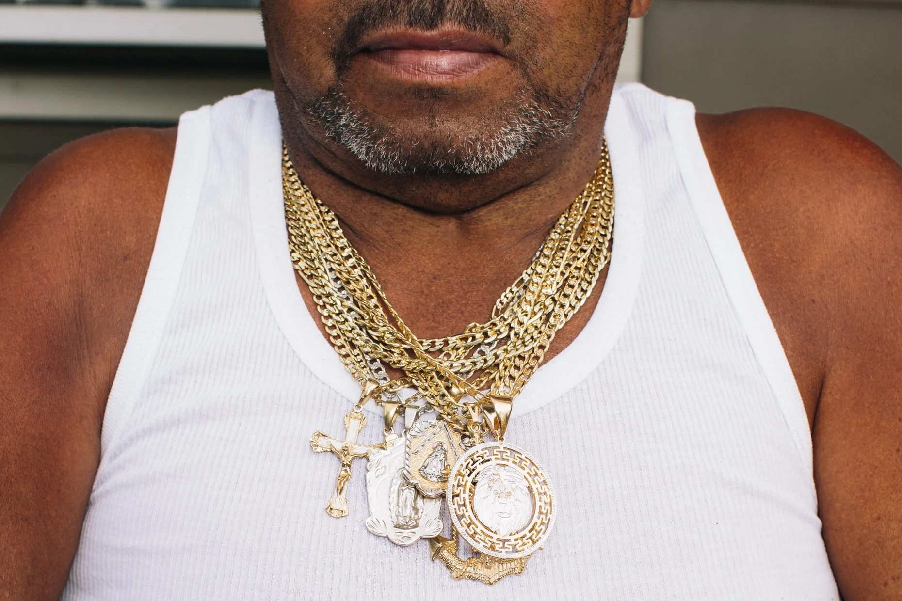 Close-up of an African American man wearing a white tank top and multiple gold chains with large, ornate pendants.