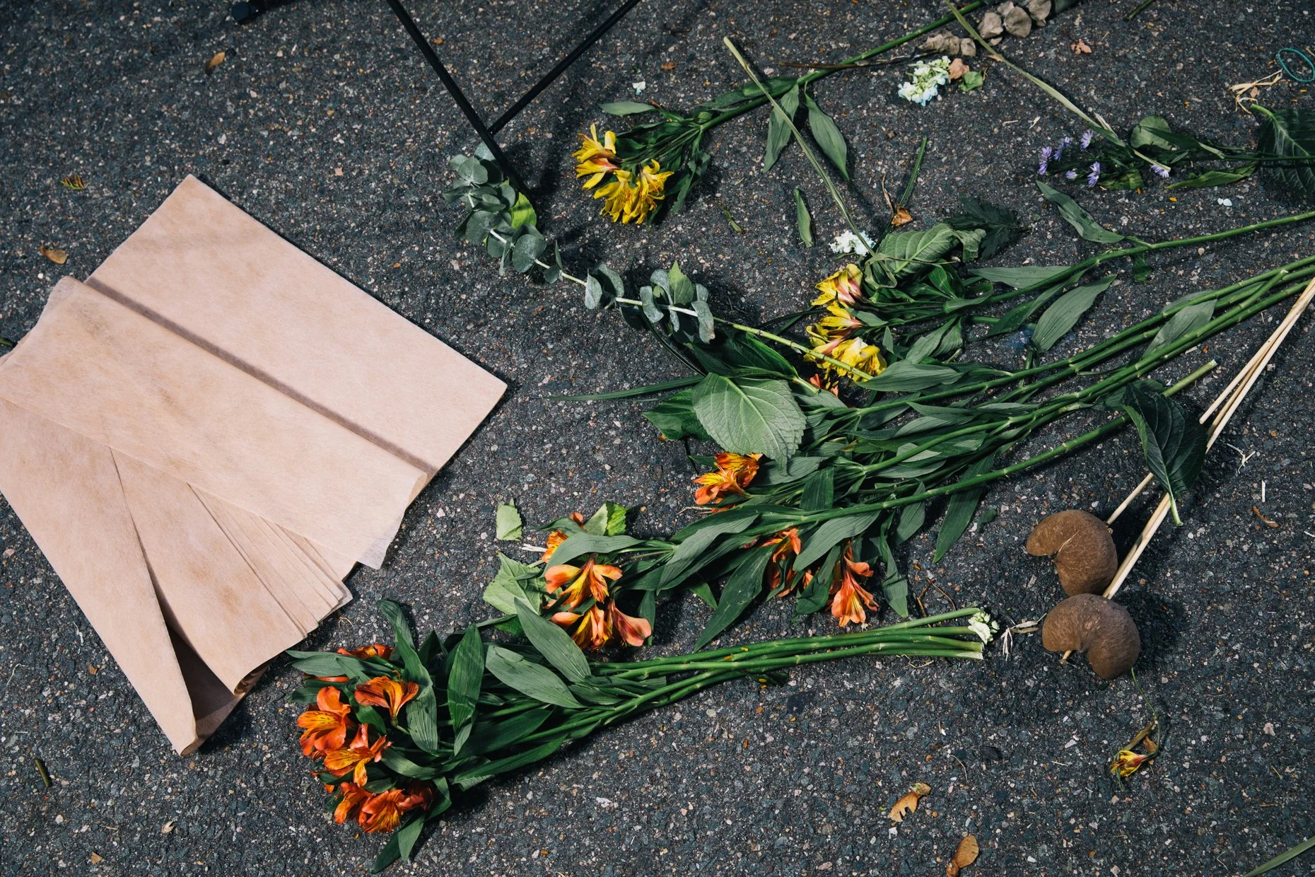 Colorful flower arrangements, some with orange and yellow lilies, are laid out on a rough asphalt road. There are paper napkins or tissues on the left side, and small rocks and scattered leaves on the ground.