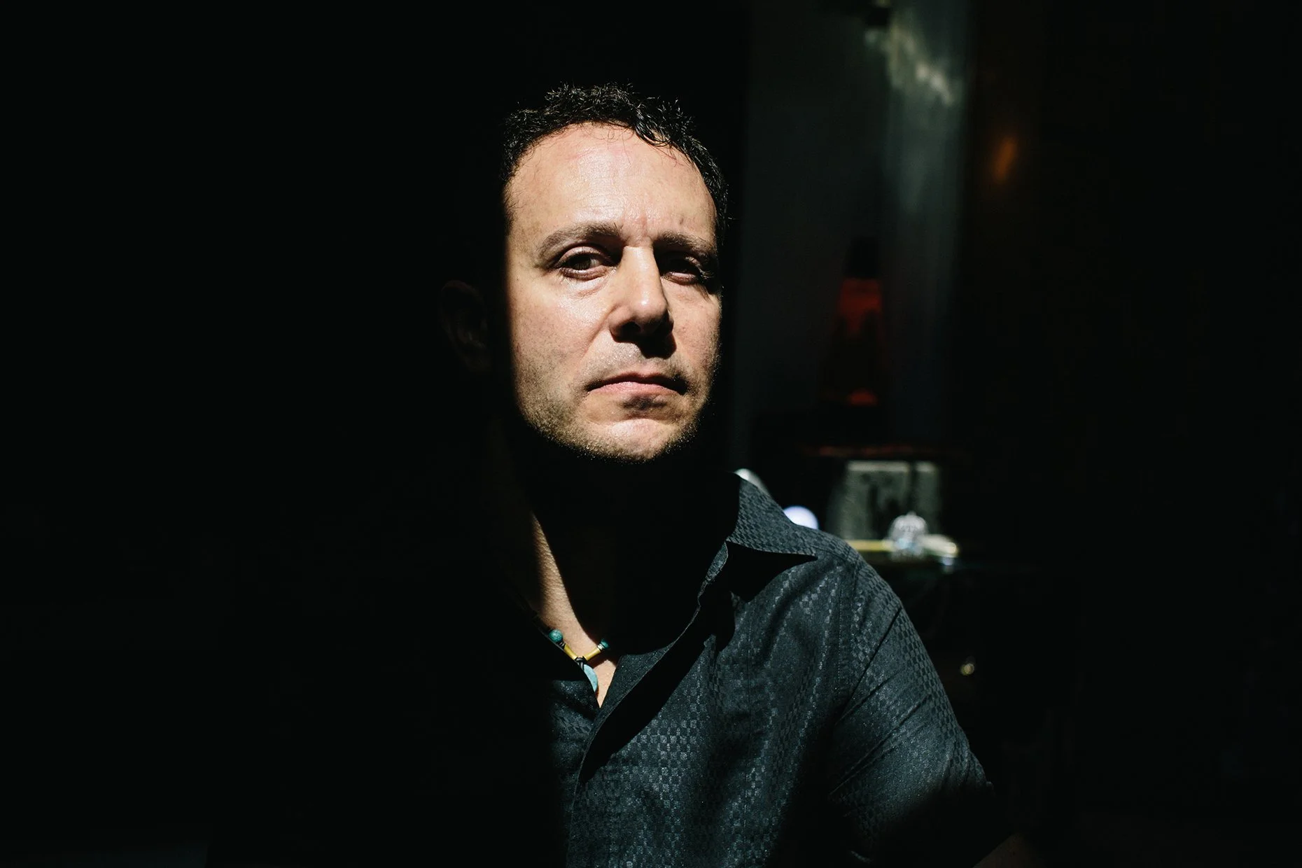 A man with short dark curly hair and light skin looking at the camera, wearing a black patterned shirt and a colorful beaded necklace, in a dimly lit environment.