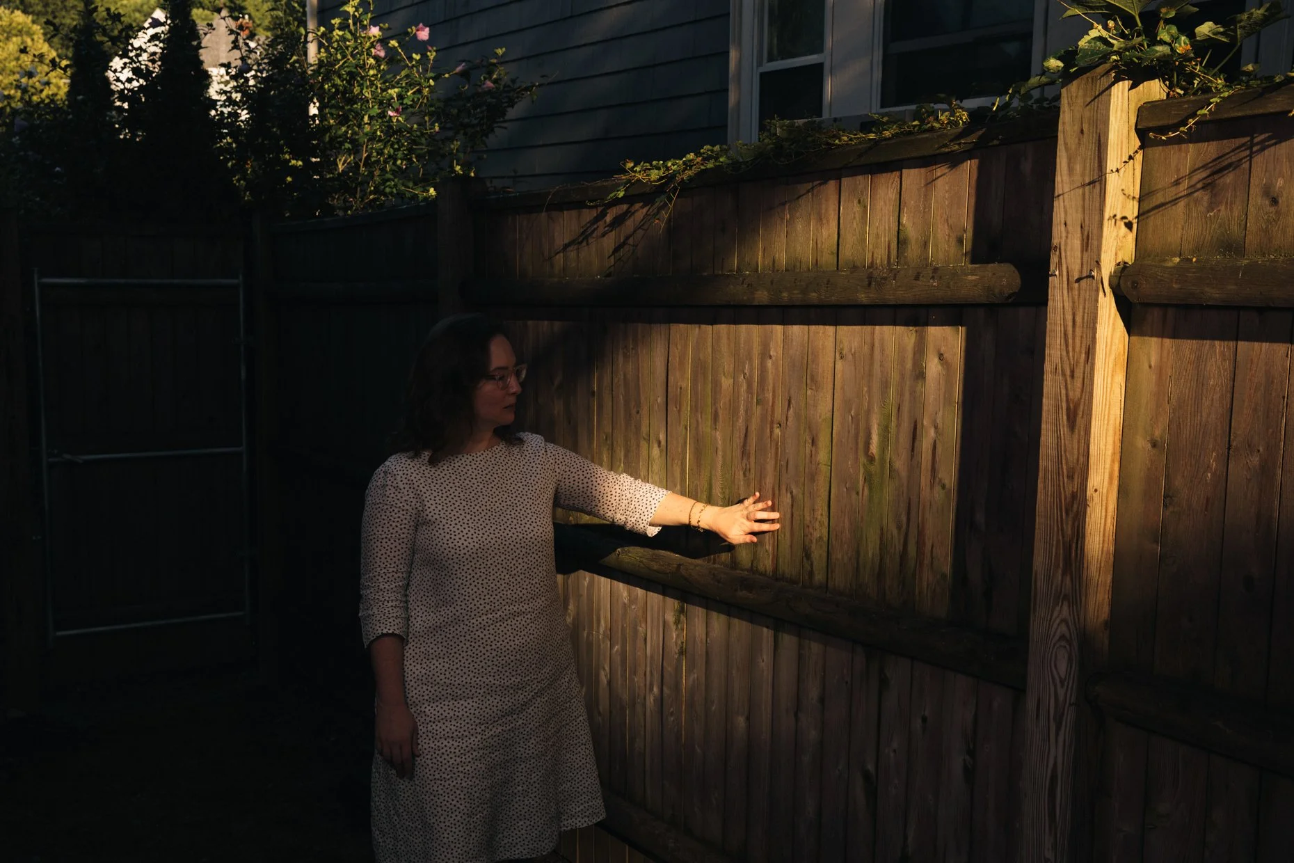A woman in a white polka dot dress with glasses standing outside at dusk, touching a wooden fence.