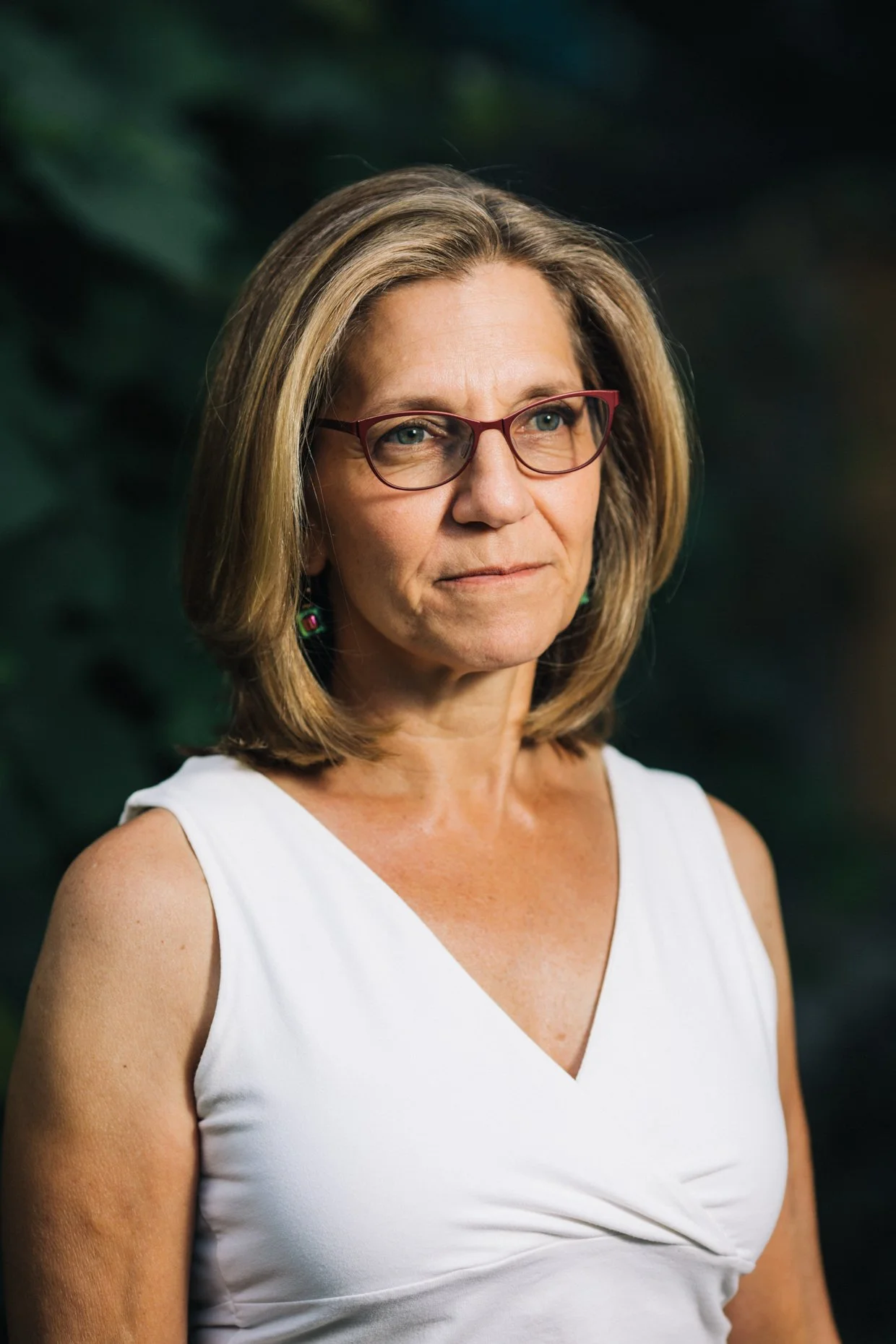 A middle-aged woman with shoulder-length blond hair, wearing red glasses, earrings, and a white sleeveless top, standing outdoors with green foliage in the background.