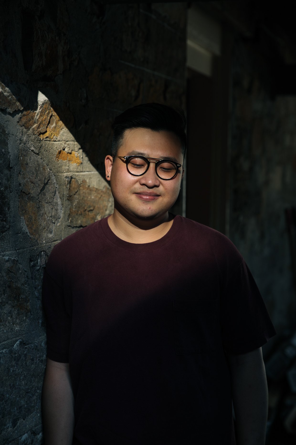 A young man with black hair, glasses, and a small earring, standing against a textured stone wall. He is wearing a dark red shirt and has a calm, content expression with eyes closed.