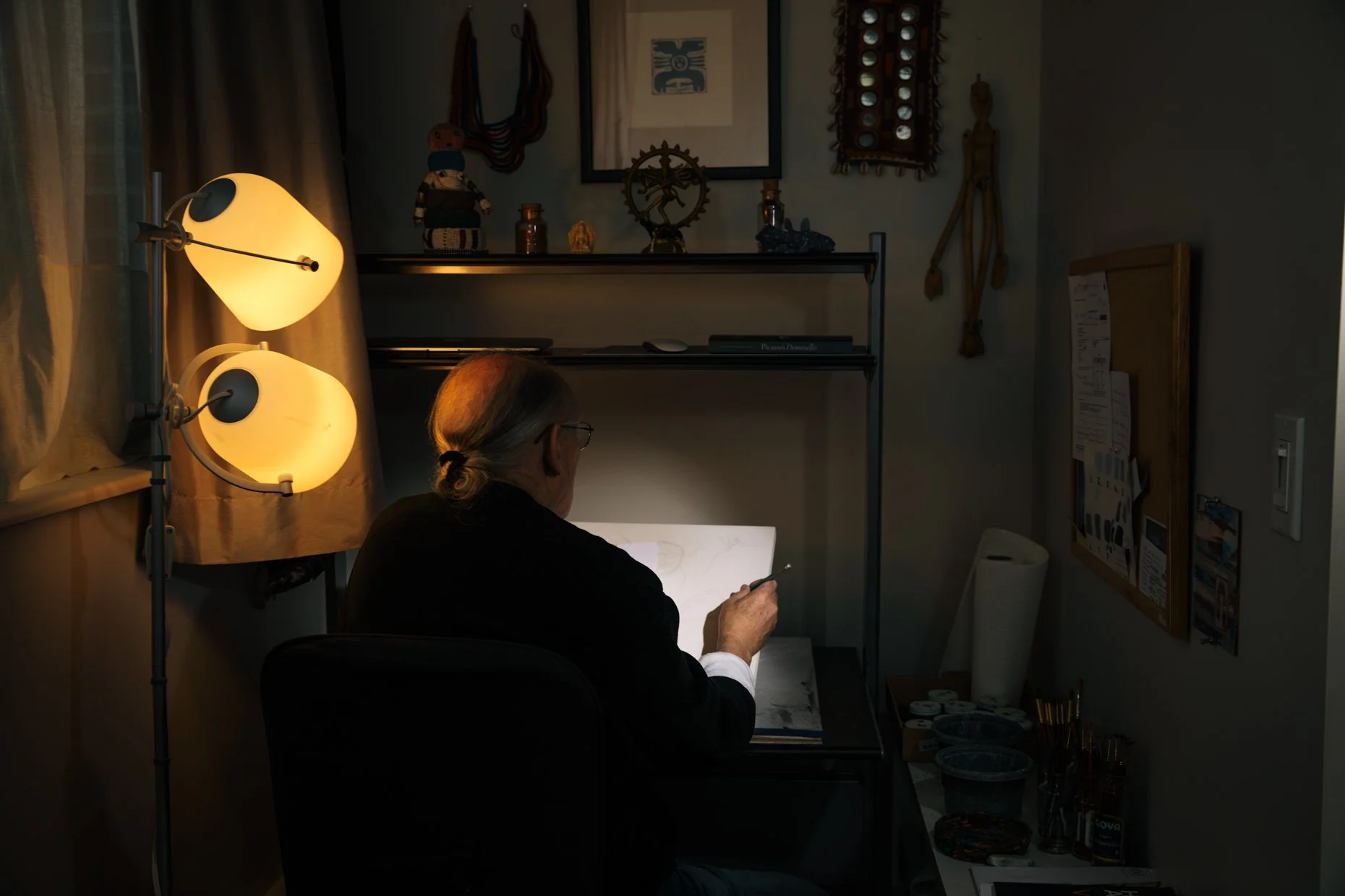 A person with gray hair tied back, wearing glasses, sitting at a desk in a dimly lit room, working on a creative project using a lightbox and drawing tools. The room has decorative items on shelves, a bulletin board, and a bright floor lamp with mult