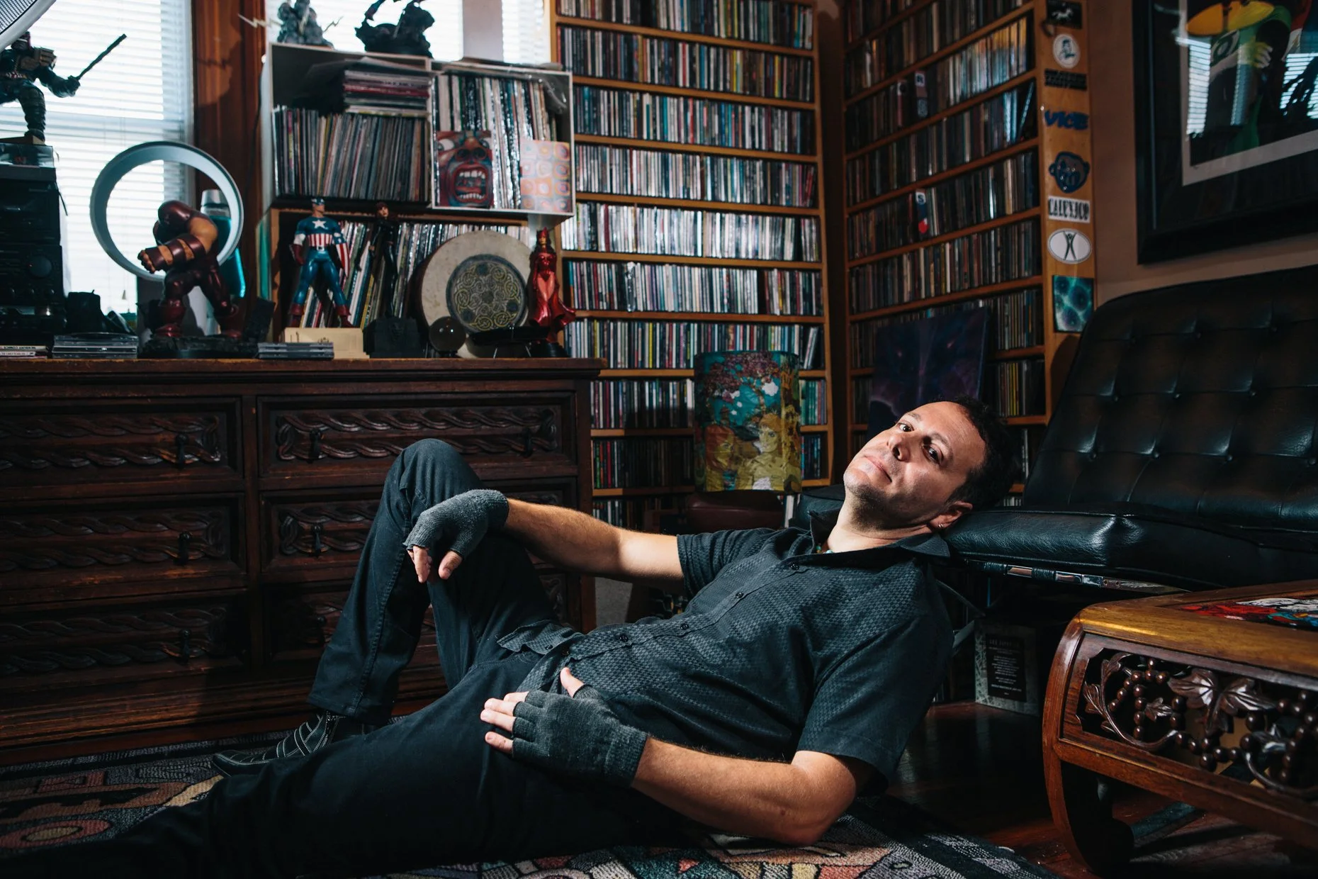 A man is reclining on a black leather chair in a room filled with books and collectibles. The room has wooden shelves with numerous books and decorative items, including superhero figures like Iron Man and Captain America, and artwork on the walls. T