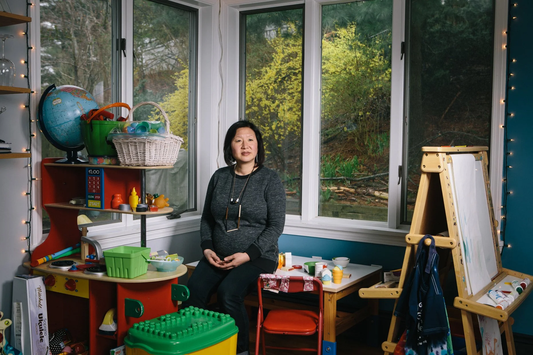 A woman sitting inside a colorful children’s playroom, with toys, a chalkboard, and large windows showing a wooded outdoor scene.