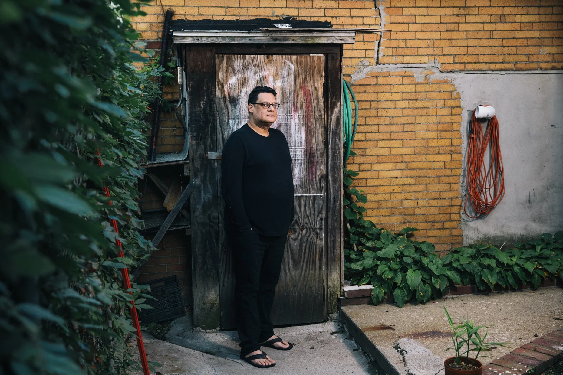 A man stands in front of a wooden shed with growth on the bottom. He wears glasses, a black long-sleeve shirt, black pants, and sandals, and is looking at the camera with a neutral expression. Surrounding plants and a brick wall are also visible.