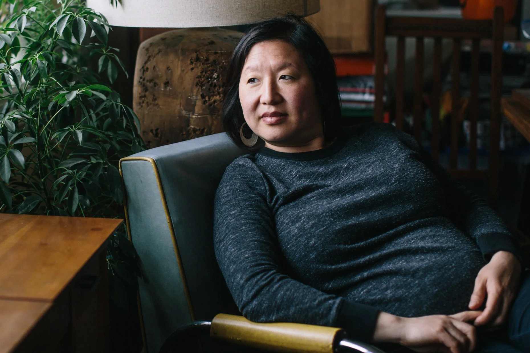 A woman with black hair and earrings sitting on a chair in a cozy indoor space with a large potted plant and wooden furniture around her.