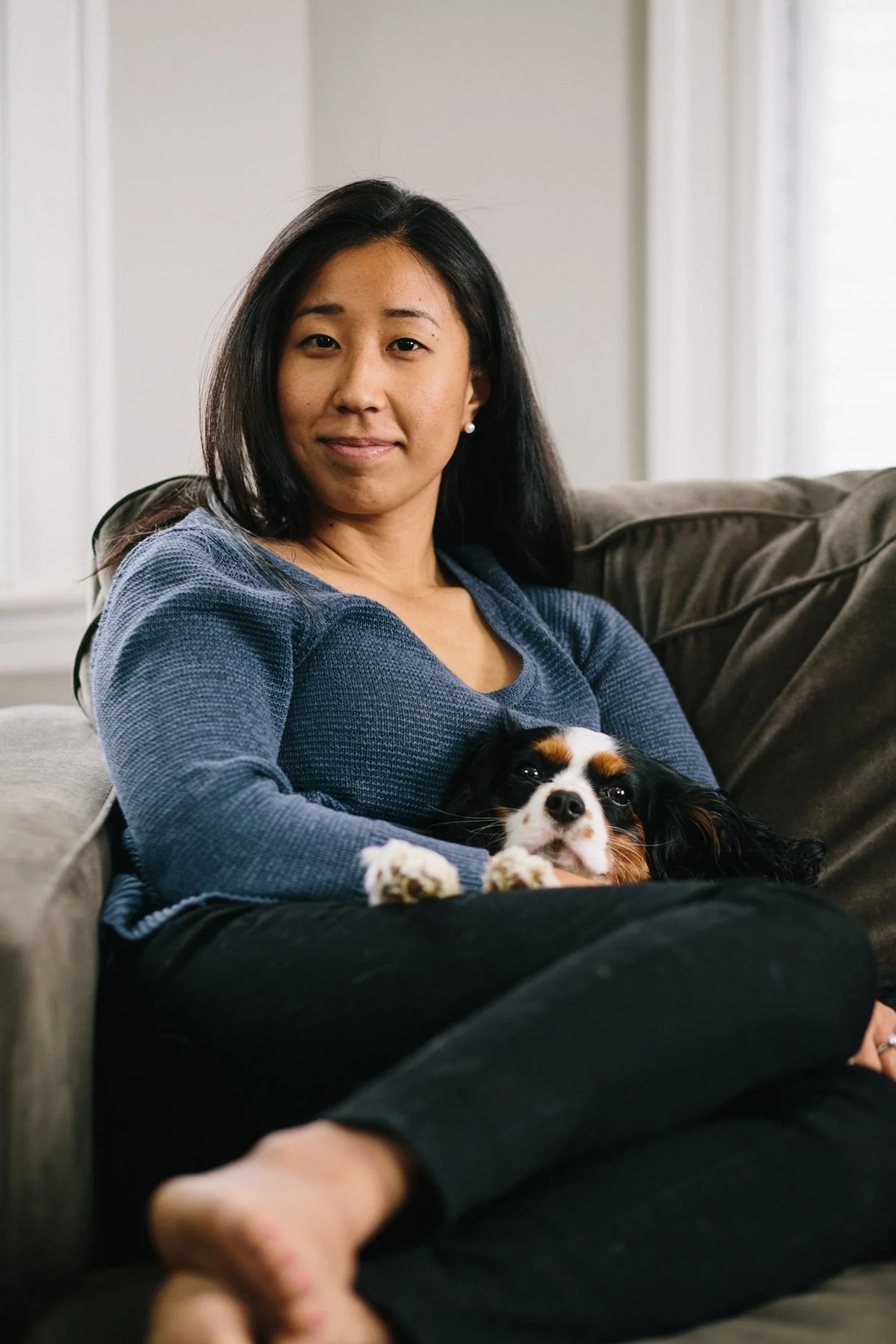 A woman with black hair and earrings sitting on a couch with a Cavalier King Charles Spaniel resting on her lap.
