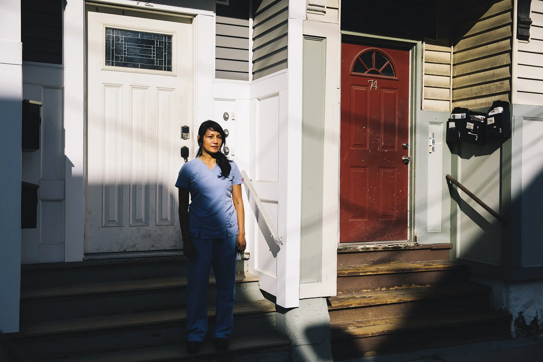 A woman in blue scrubs standing in front of two house entrances, one with a white door and the other with a red door, daytime.