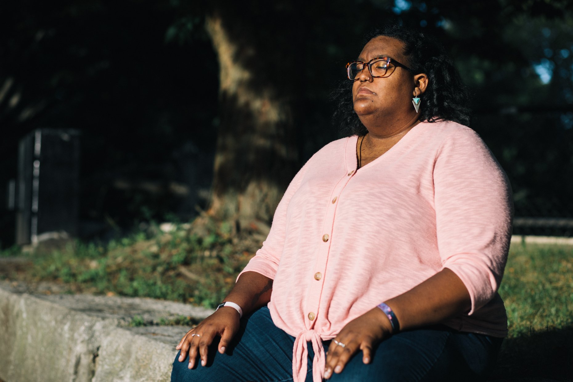A woman with glasses and earrings sitting outdoors, wearing a light pink button-up shirt, with her eyes closed and hands resting on her lap, in a park-like setting with trees and greenery in the background.