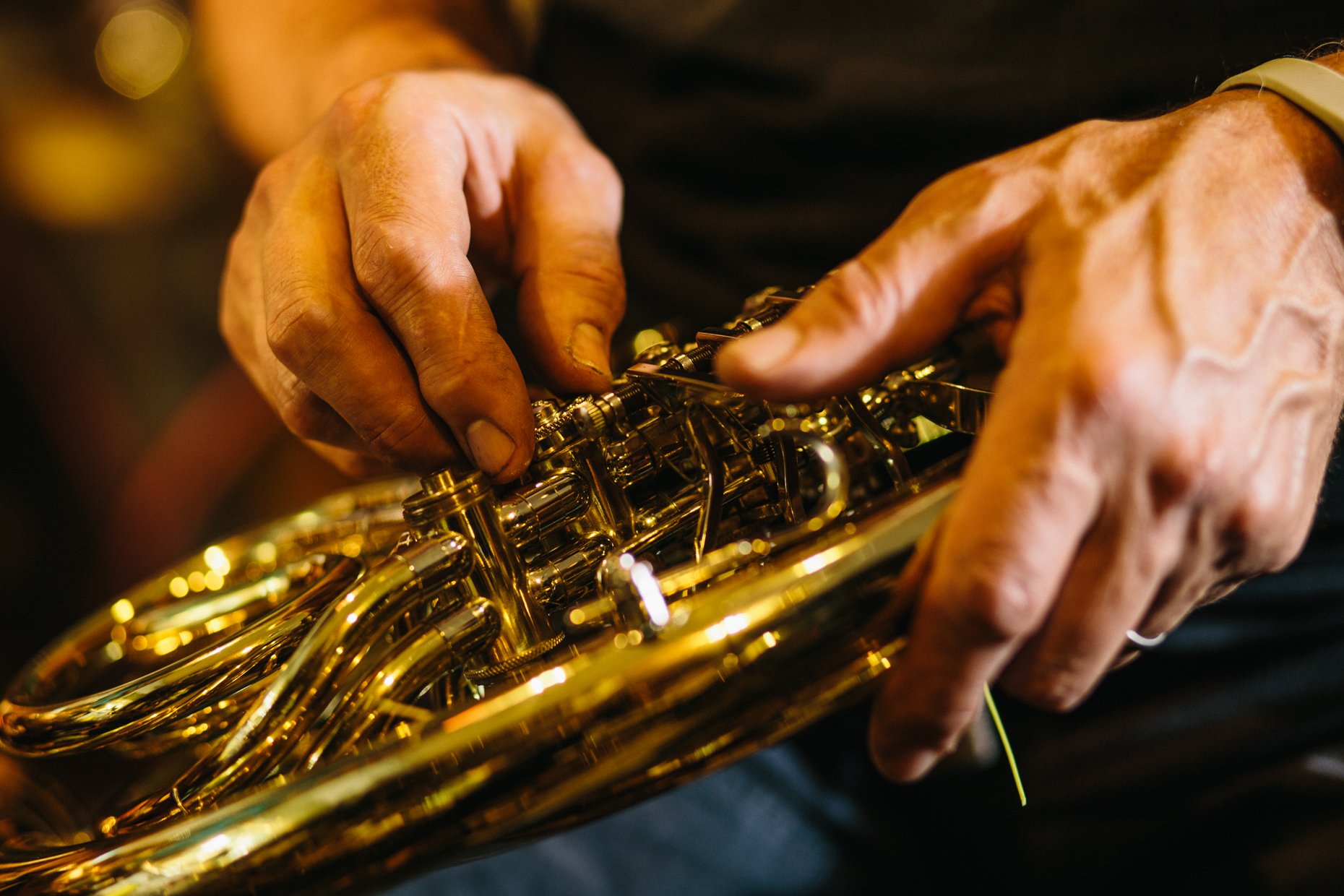 Close-up of a person’s hands adjusting a shiny, gold-colored wind instrument, likely a saxophone, with blurred background.