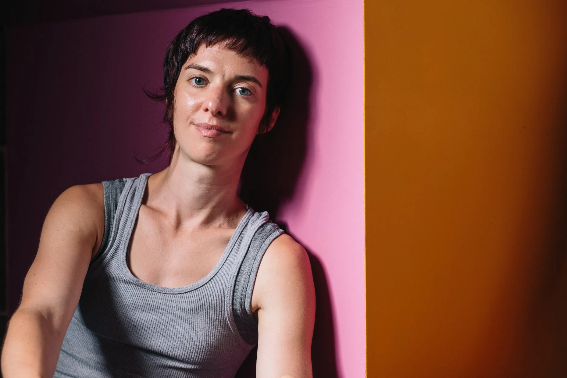 A woman with short dark hair and blue eyes, wearing a gray sleeveless top, leaning against a pink wall with a brown wall on the right side.