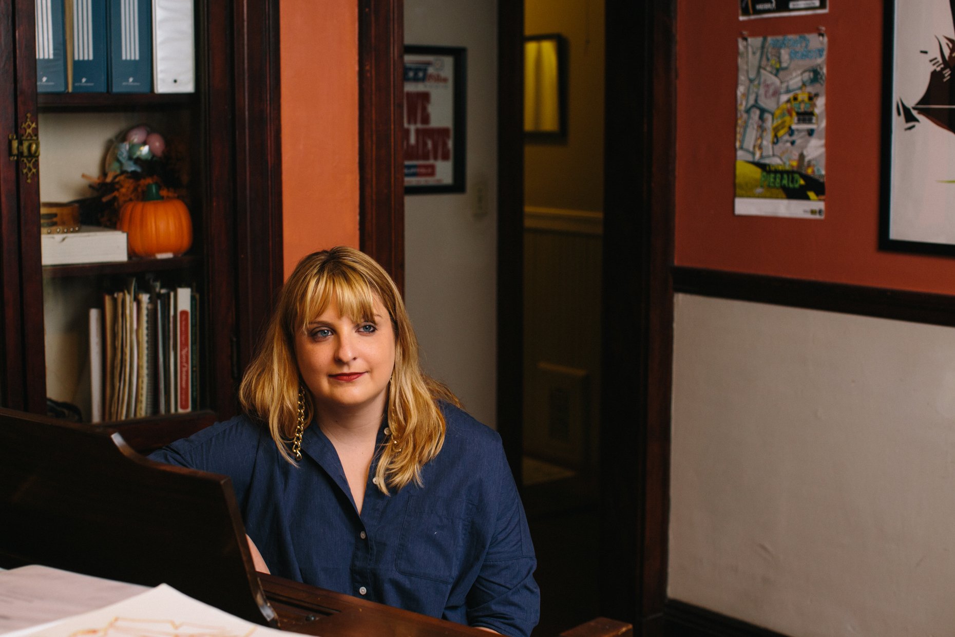 A woman with blonde hair, blue eyes, wearing a navy blue shirt and a gold chain, sitting at a piano in a room decorated with framed artwork and shelves with books and a pumpkin.