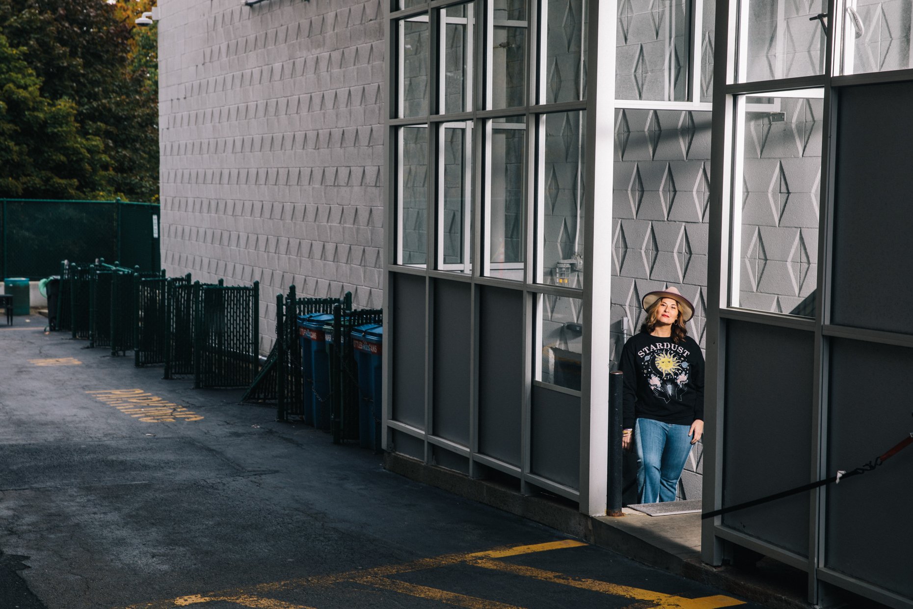A woman stands at a corner outside a building, partially inside a glass-enclosed structure. She wears a wide-brimmed hat and a black sweatshirt with a celestial graphic that says 'Stardust.' The building has a textured gray wall with a pattern of pro