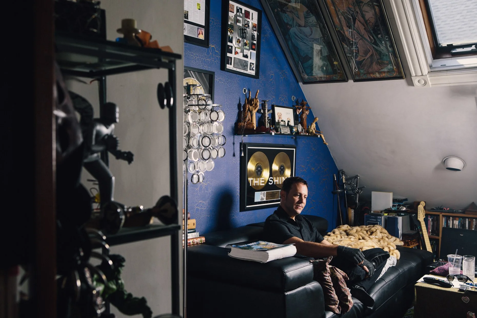 A man sitting on a black leather couch in a room with a blue textured wall, decorated with a gold record and various framed pictures and artwork. The room has a sloped ceiling with a window and various collectibles, trophies, and books on shelves. Th