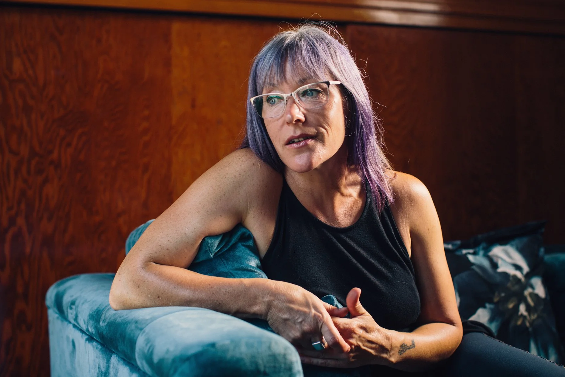 A woman with purple-tinted hair, glasses, and a black sleeveless top sitting on a blue velvet couch against a wooden paneled wall.